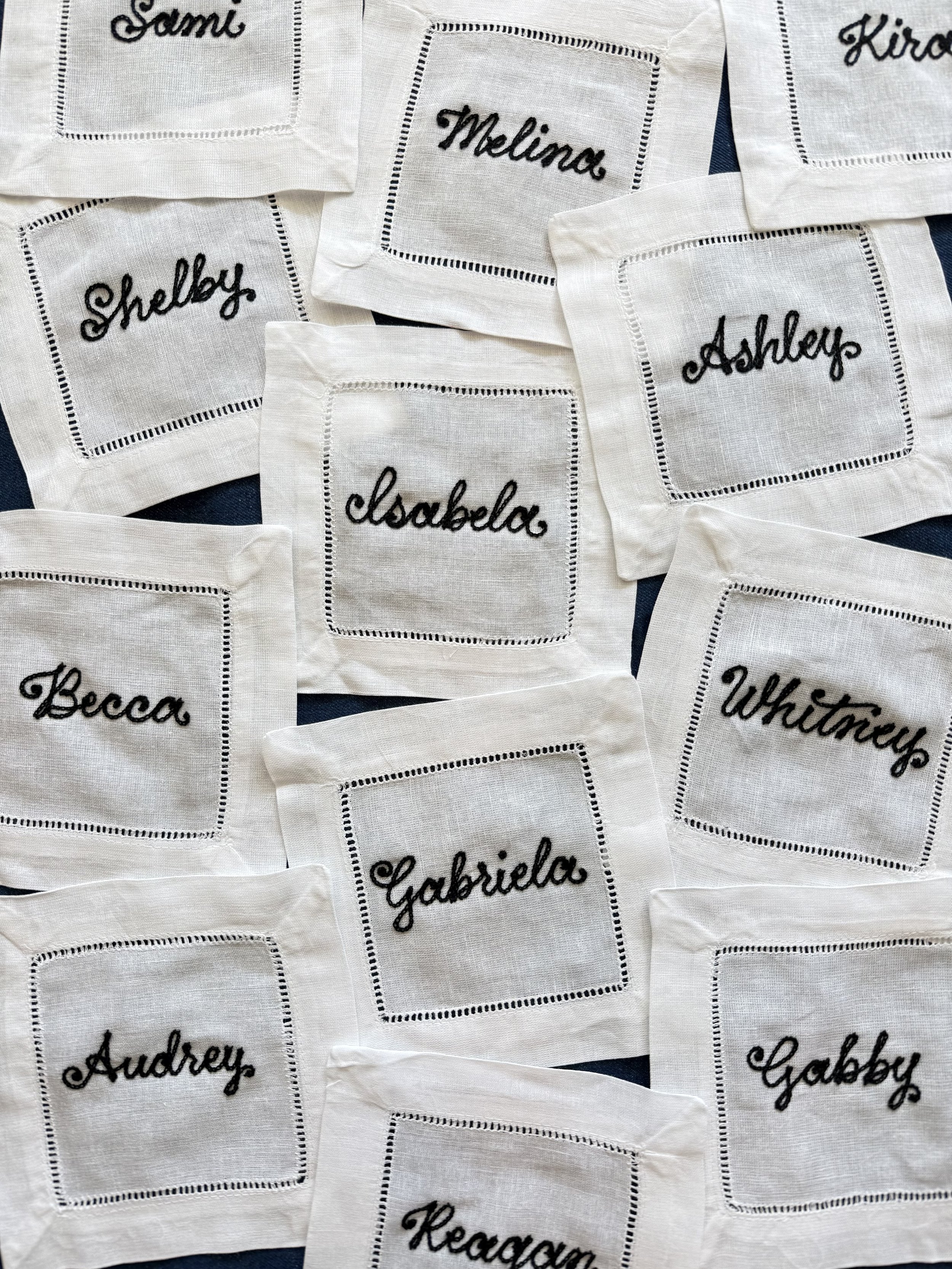 Multiple white cocktail napkins with chainstitch embroidered black cursive names such as Shelby, Melina, Ashley, Gabriela, Becca, Audrey, Reagan, and Whitney. Premium gifting for influencer dinner in Fort Worth, Texas.