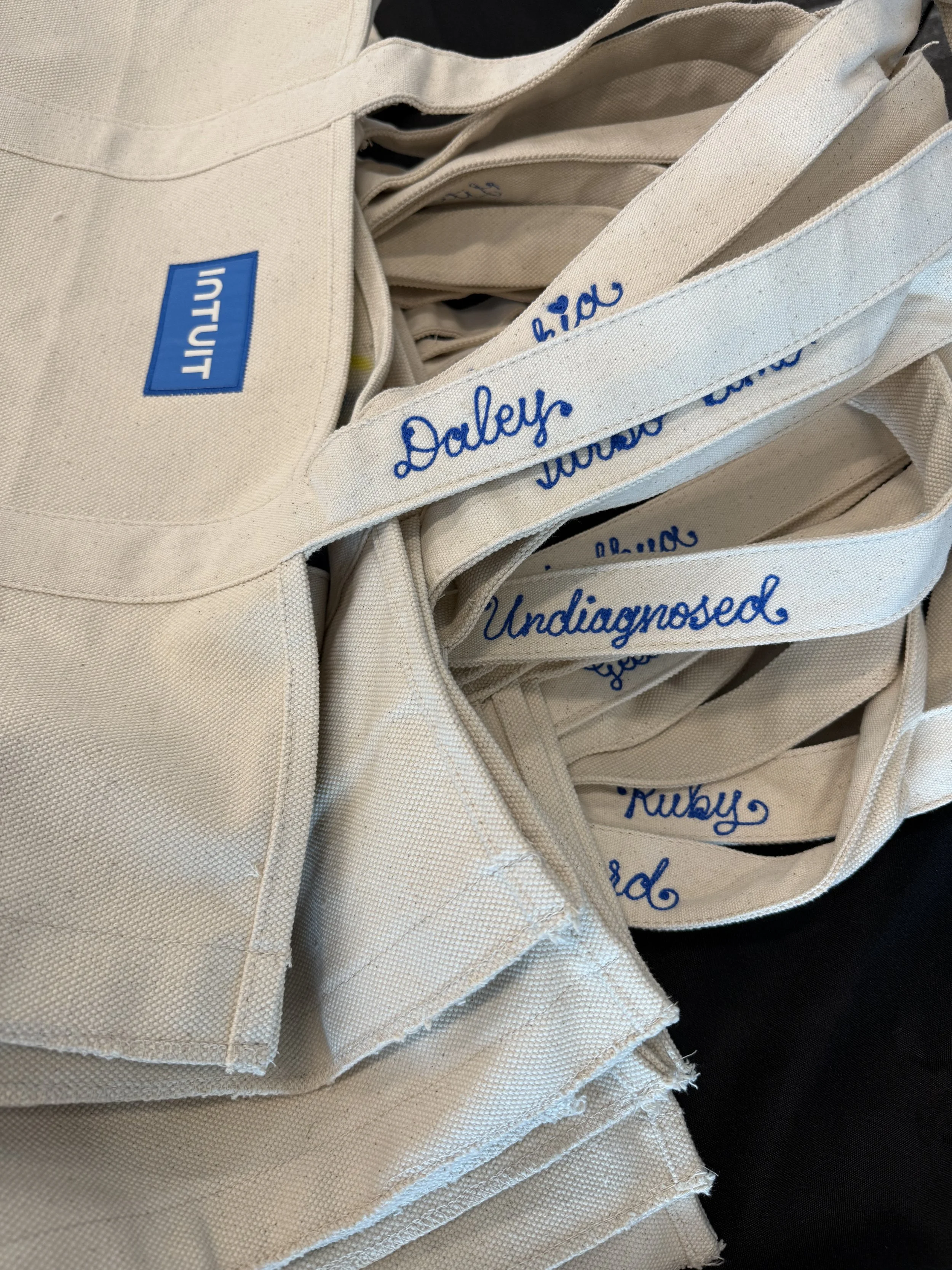 Multiple beige tote bags with blue chainstitch embroidered names such as "Undiagnosed," "Darcy," and other names. Double Vision was on-site in Charlotte, North Carolina to provide live embroidery for Intuit employees.