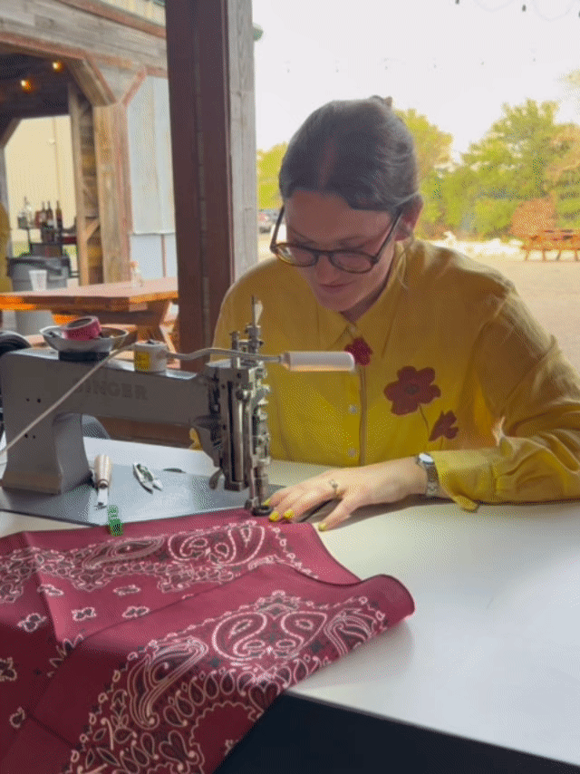 Woman with dark hair and glasses sewing a red bandana on a sewing machine, indoors with a view of trees outside.