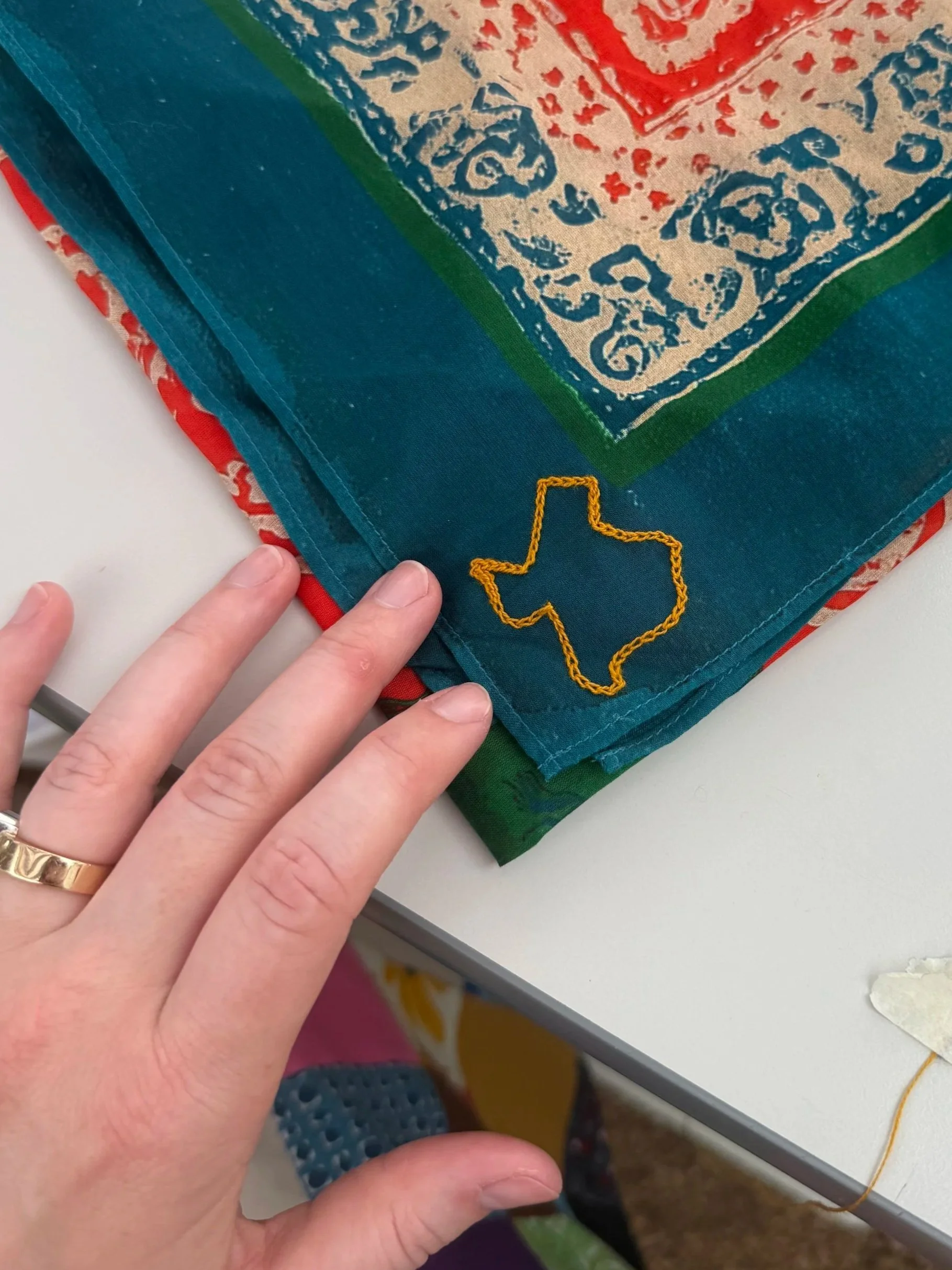 Close-up of a hand with a gold ring holding the corner of a colorful fabric with a chainstitch embroidered outline of Texas in yellow thread. In Round Top, Texas.
