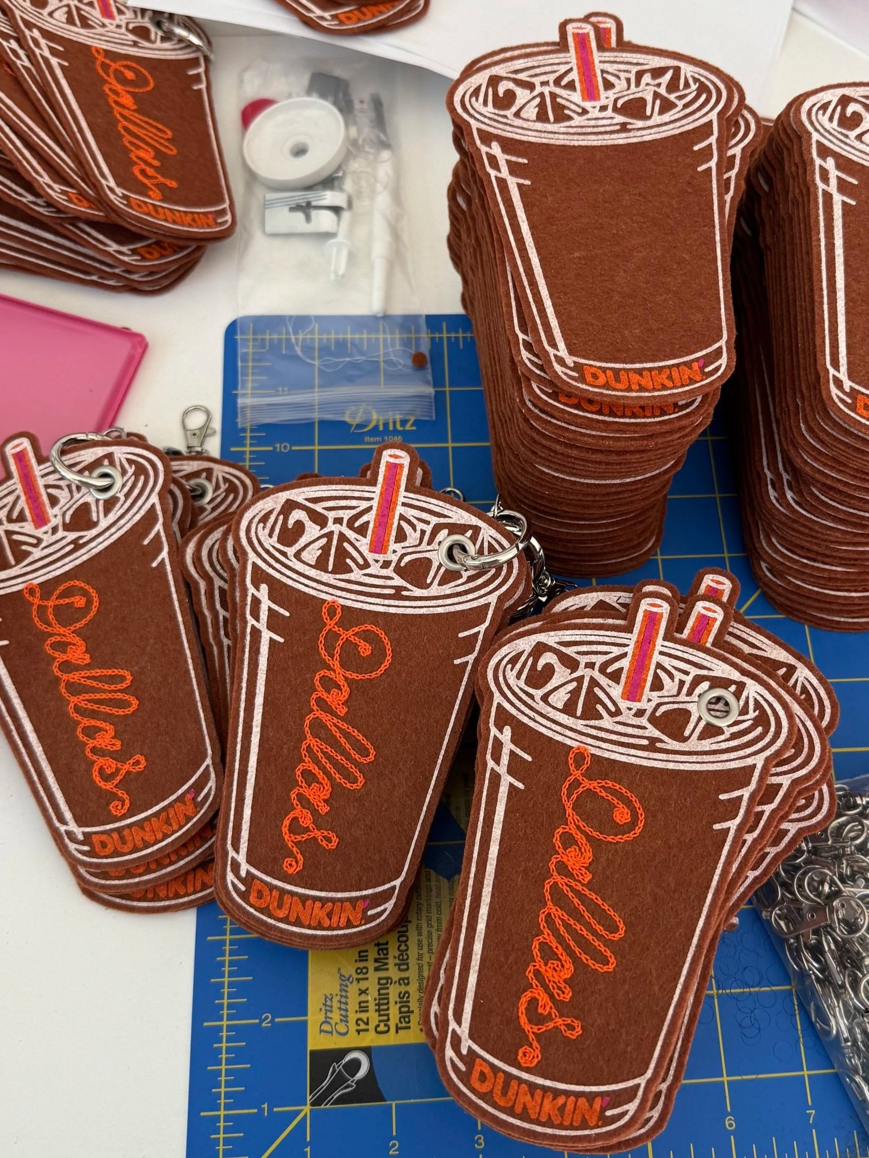 Stack of brown felt keychains shaped like Dunkin' iced coffee cups with a pink straw, featuring the words 'Dallas' in orange embroidery, placed on a cutting mat with crafting tools around.