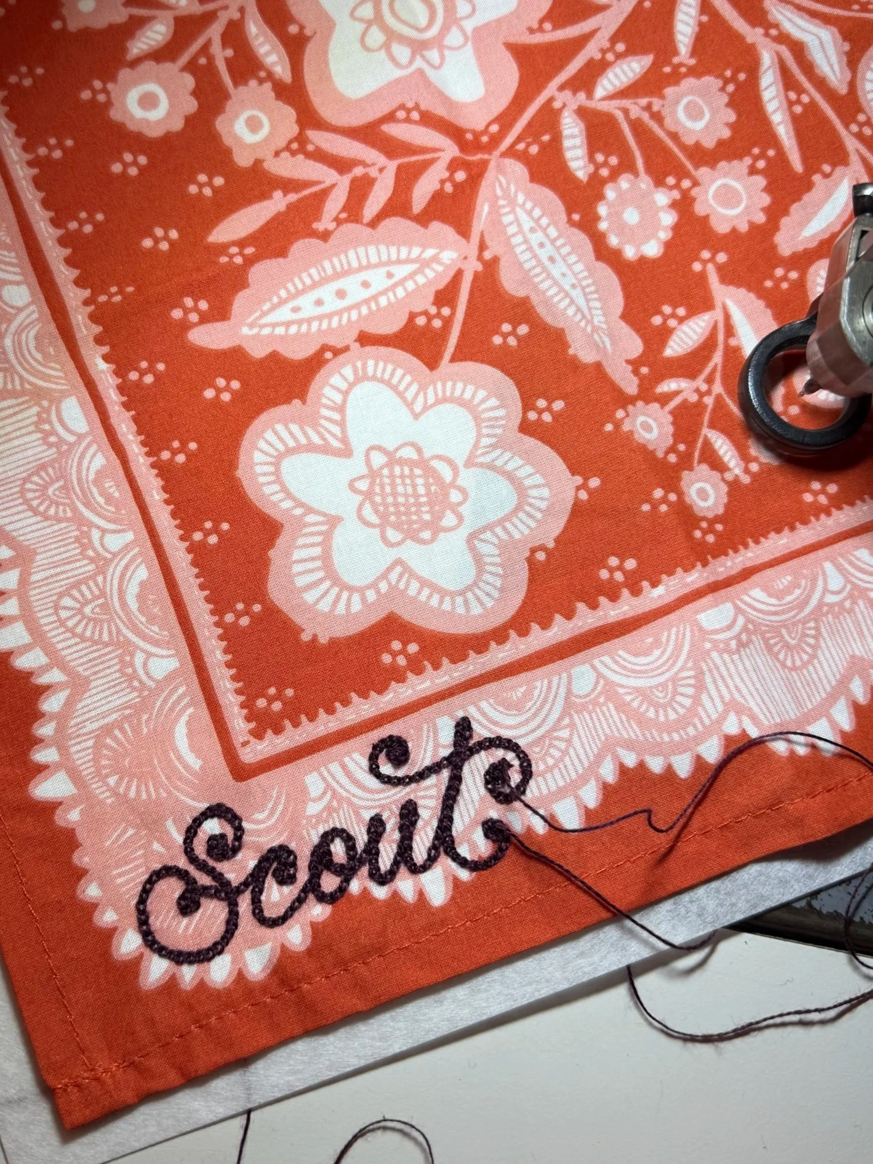 Close-up of an orange bandana with white floral patterns and the embroidered word 'Scrubs' in black thread.
