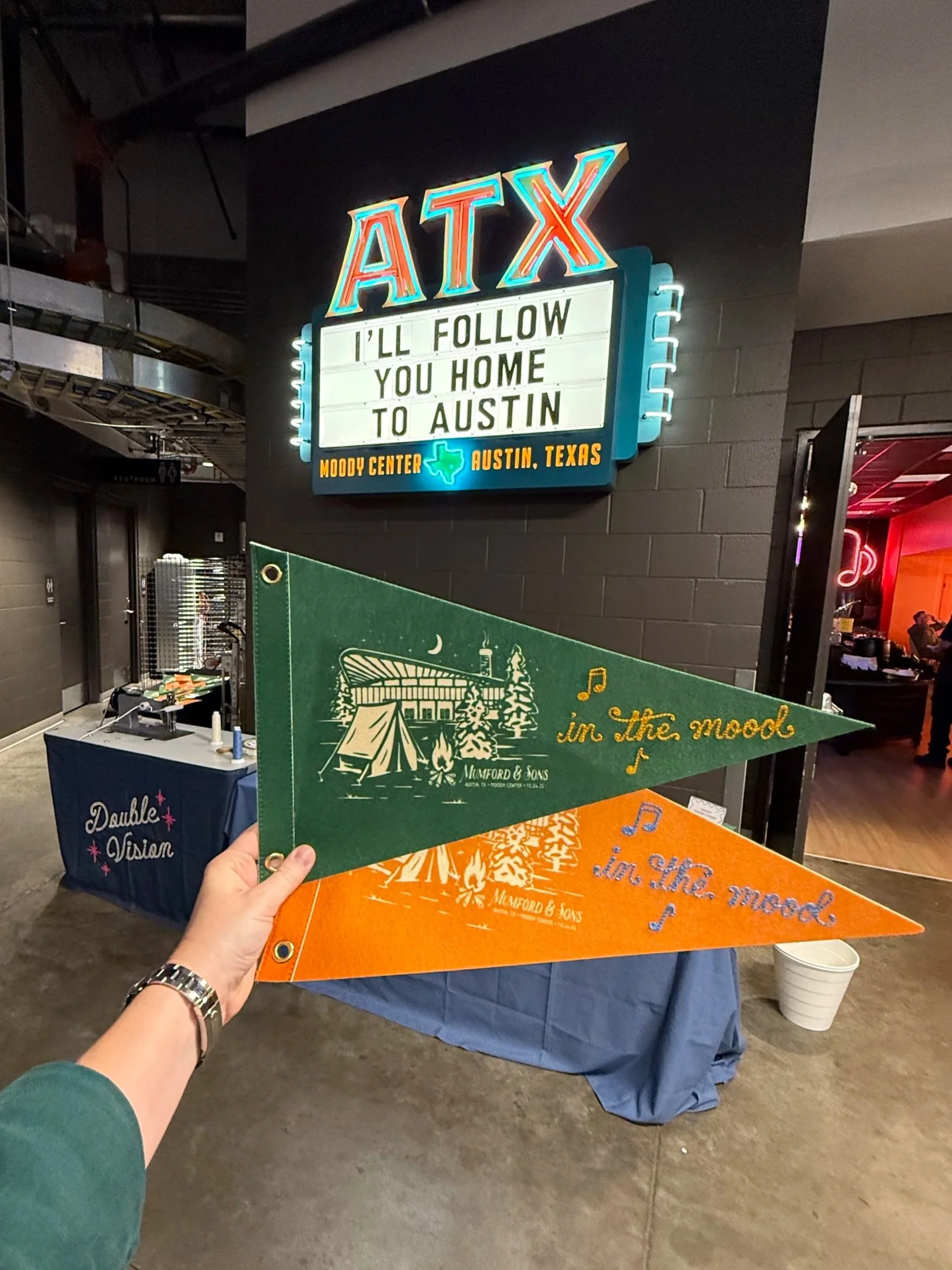 Person holding green and orange pennants with 'in the mood' chainstitched on them, against a backdrop of a neon sign reading 'ATX' and a marquee sign with the message 'I'll follow you home to Austin', at Moody Center in Austin, Texas.
