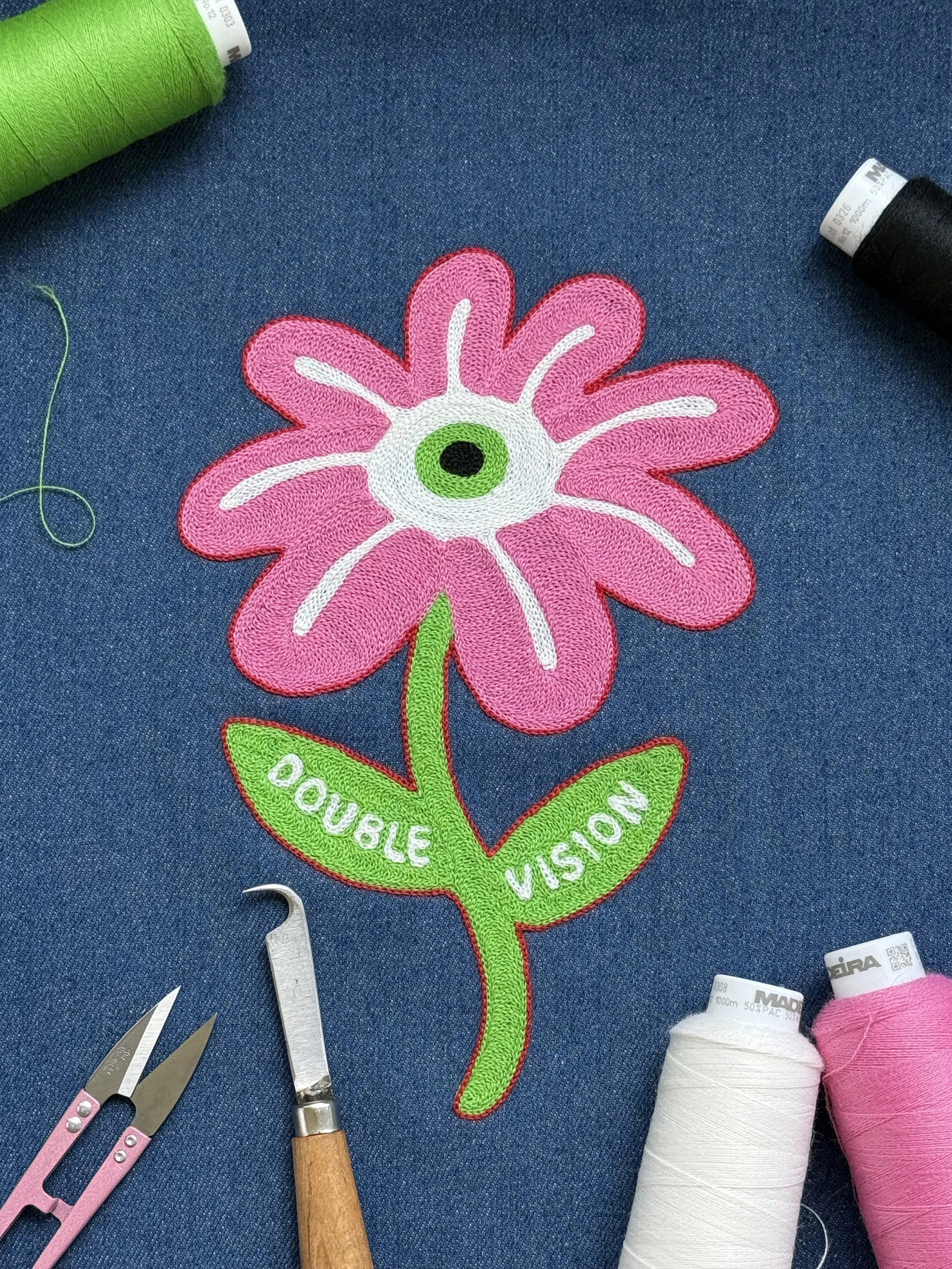 A colorful embroidered patch of a flower with pink petals, a green and black center, and green leaves with the words "Double Vision" on the leaves, laid out on a blue fabric background alongside sewing tools and thread spools.