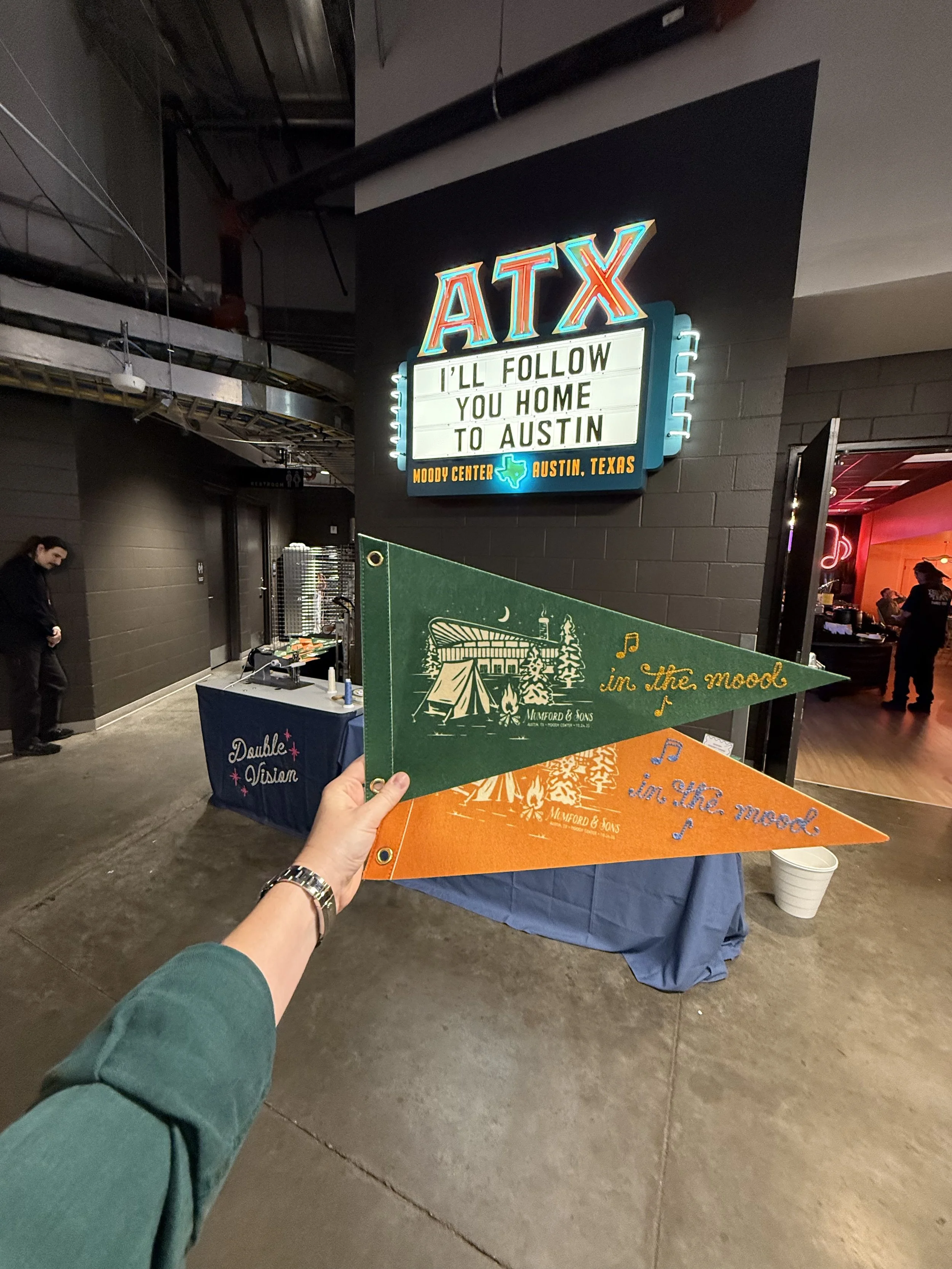 Person holding green and orange pennants with 'in the mood' chainstitched on them, against a backdrop of a neon sign reading 'ATX' and a marquee sign with the message 'I'll follow you home to Austin', at Moody Center in Austin, Texas.