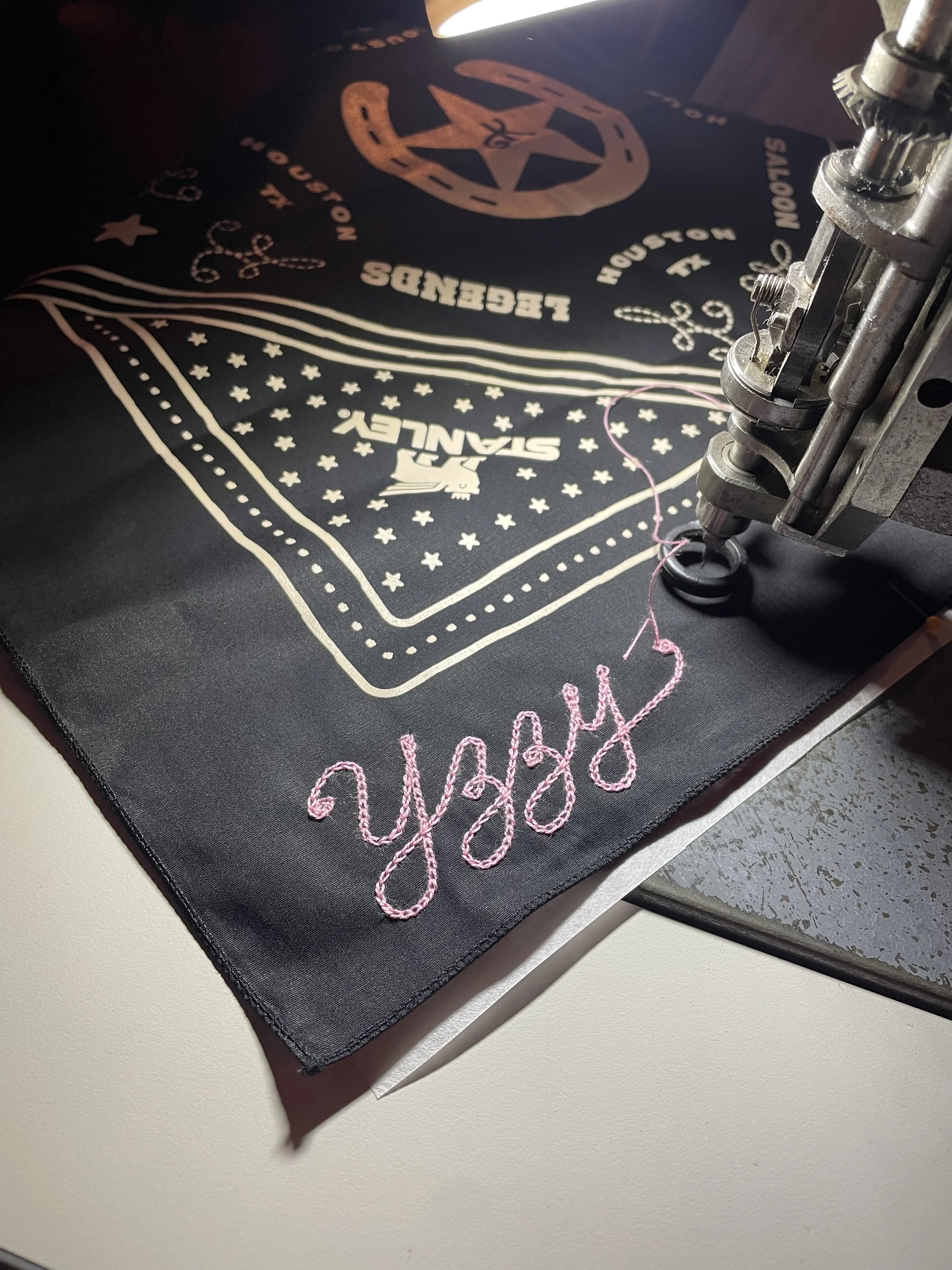 A chainstitch embroidery machine stitching the word 'Yzzy' in pink thread onto black fabric with a design featuring a bandana pattern. Double Vision was on-site to provide live chainstitch for Stanley 1913 x Barbie collab event in Houston, Texas.