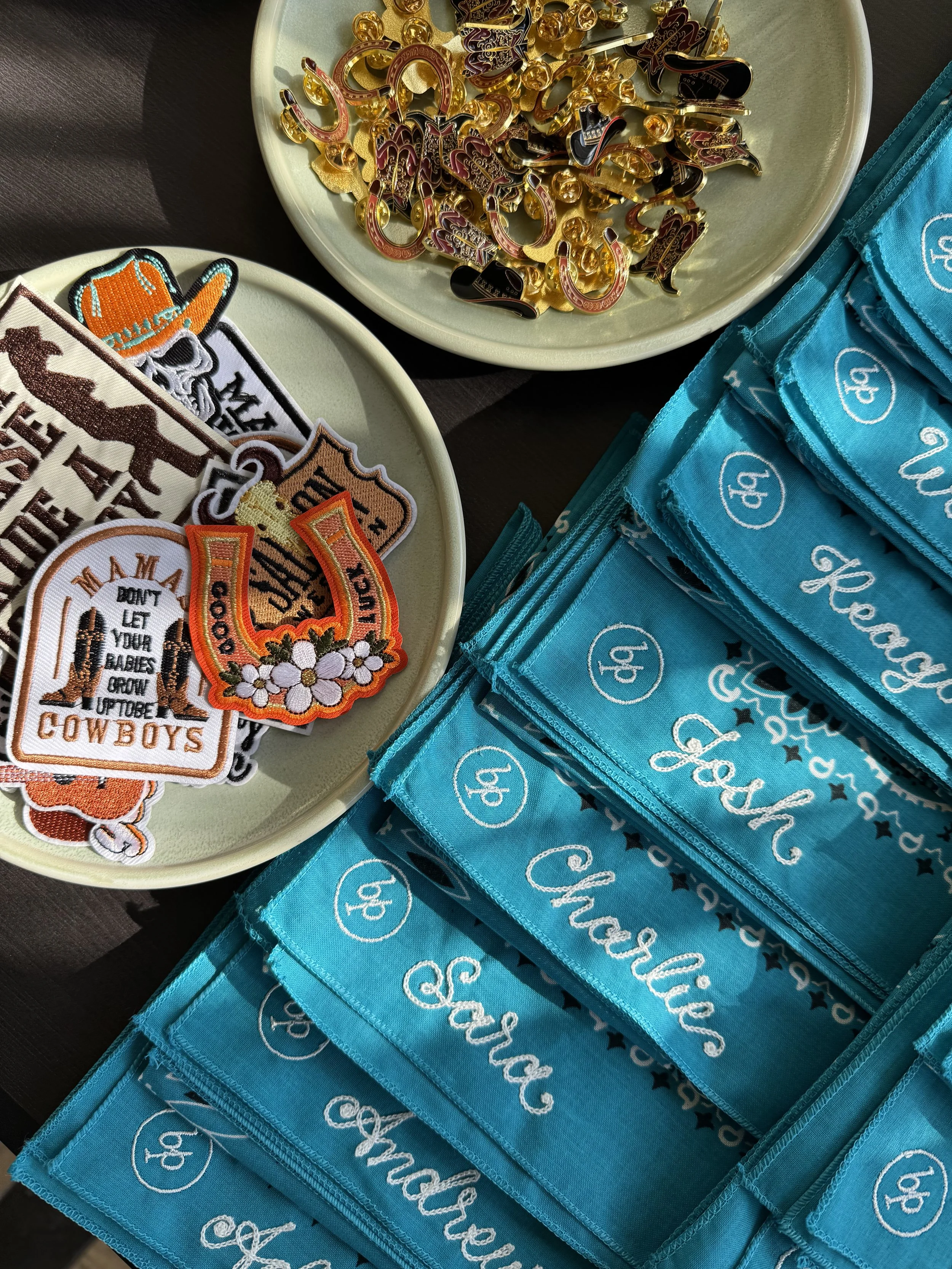 Two plates of embroidered patches with cowboy and western themes, and a stack of blue fabric pouches with white cursive text reading 'Joy, Peace, Faith, Love, Share, Faith' and decorative patterns.