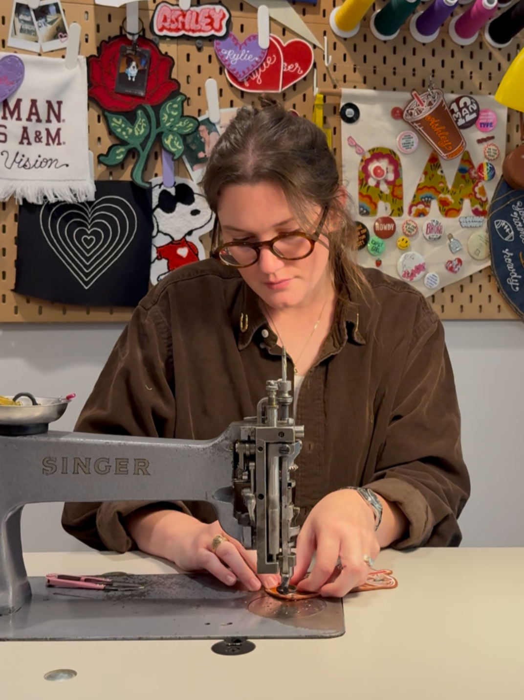 A woman sewing on a vintage Singer chainstitch machine at a desk, with colorful patchwork and decorative items on a pegboard behind her.