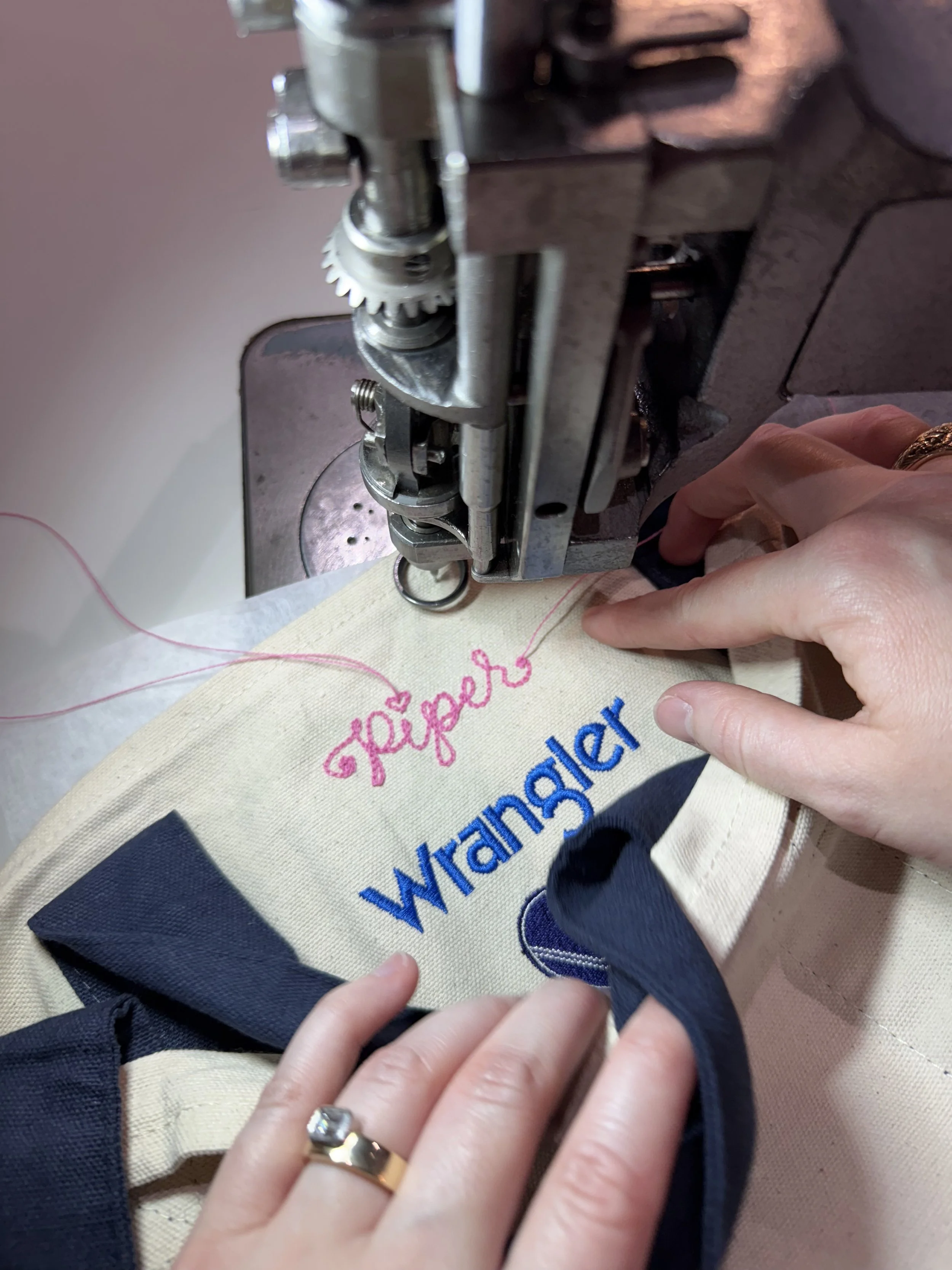 Chainstitch machine stitching a mini tote bag with the words 'Wangler' in blue and 'Piper' in pink, with hands guiding the fabric. Double Vision was in store to provide live chainstitch embroidery for Wranger customers in Waco, Texas.