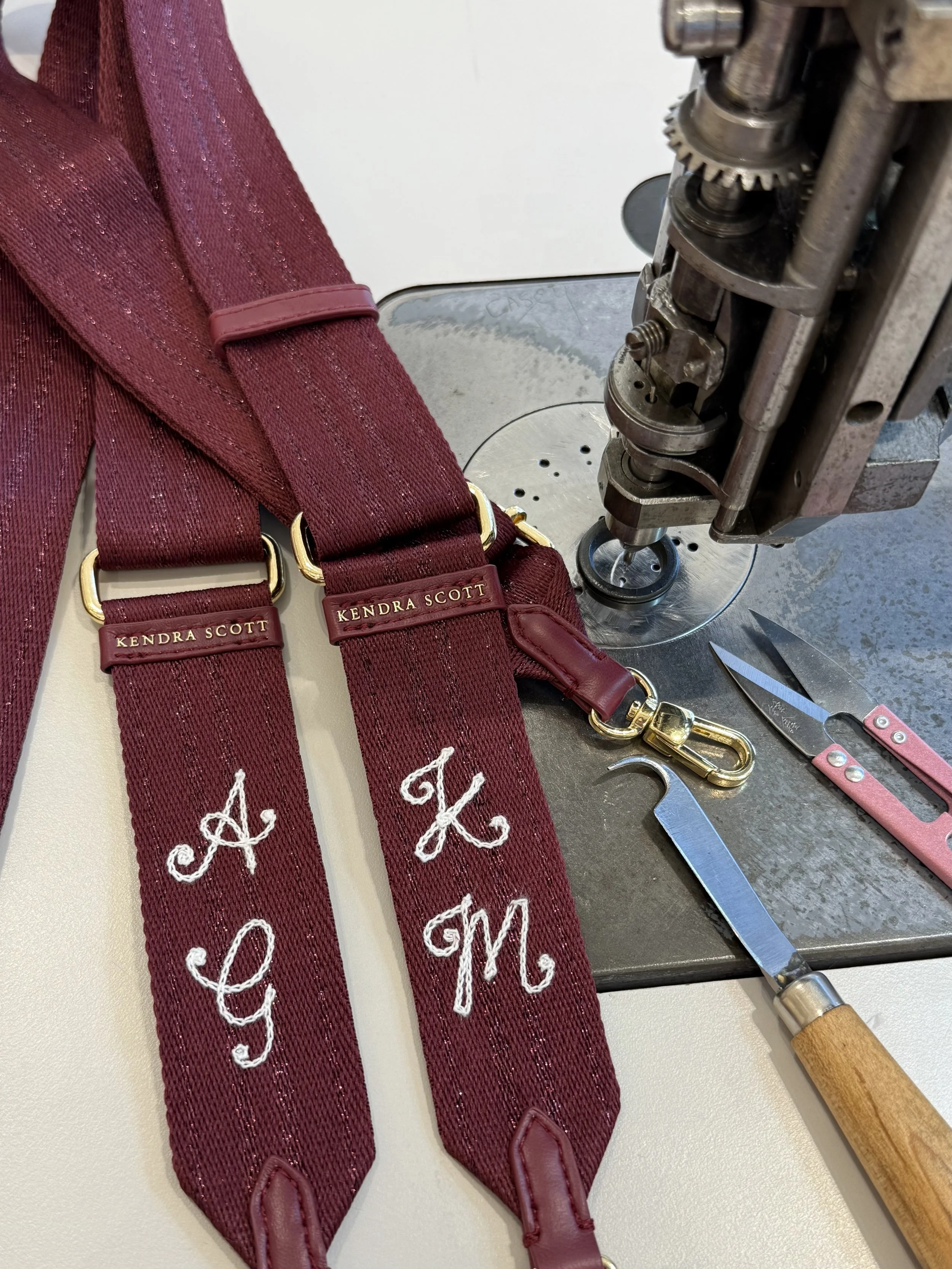 Close-up of maroon bag strap with chainstitch embroidered white initials, labeling, and hardware, placed on a worktable next to sewing and cutting tools, with a chainstitch machine in the background.