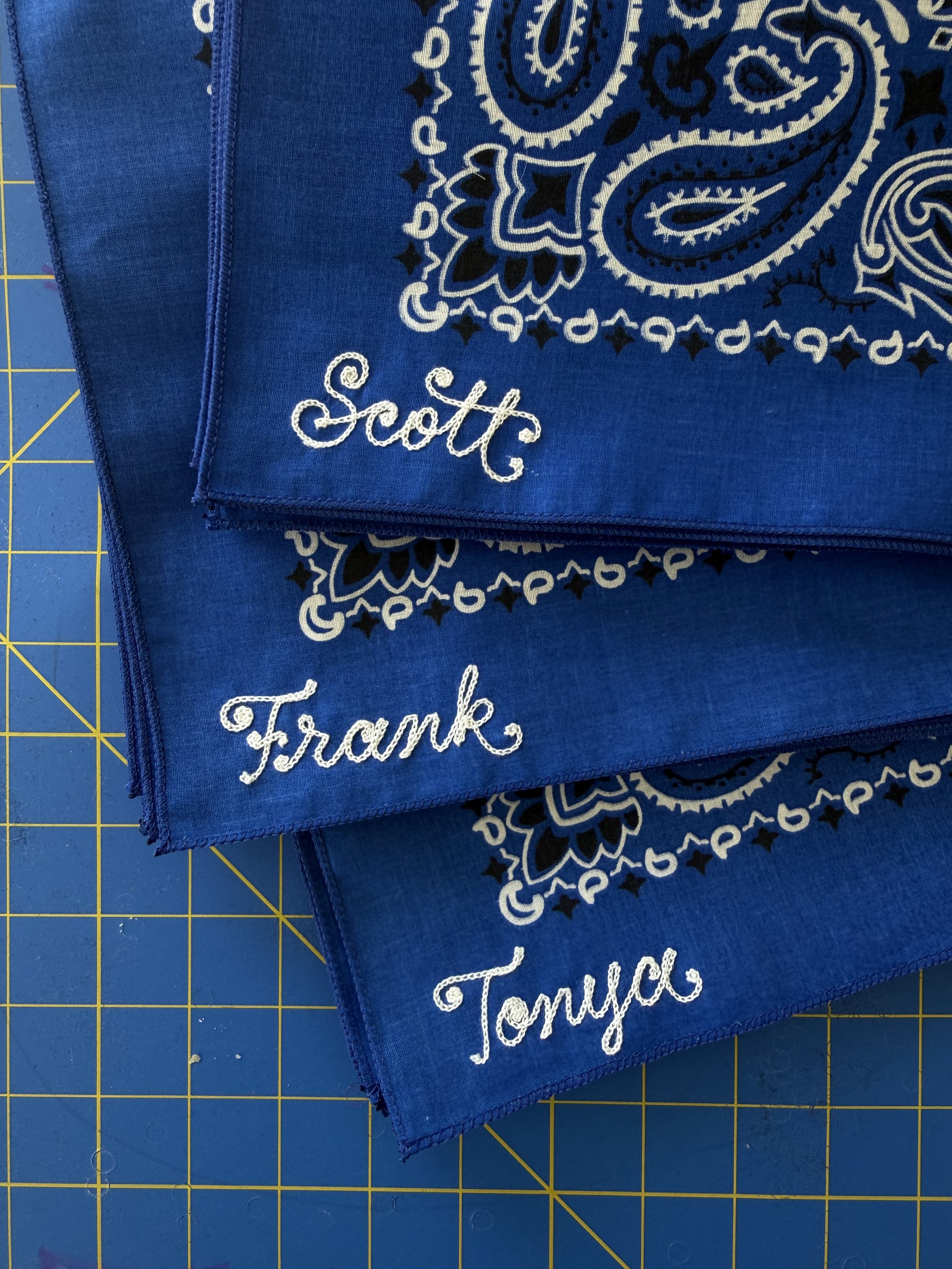 Royal blue bandanas with white chainstitch embroidered names: Scott, Frank, and Tonya, placed on a yellow grid cutting mat.