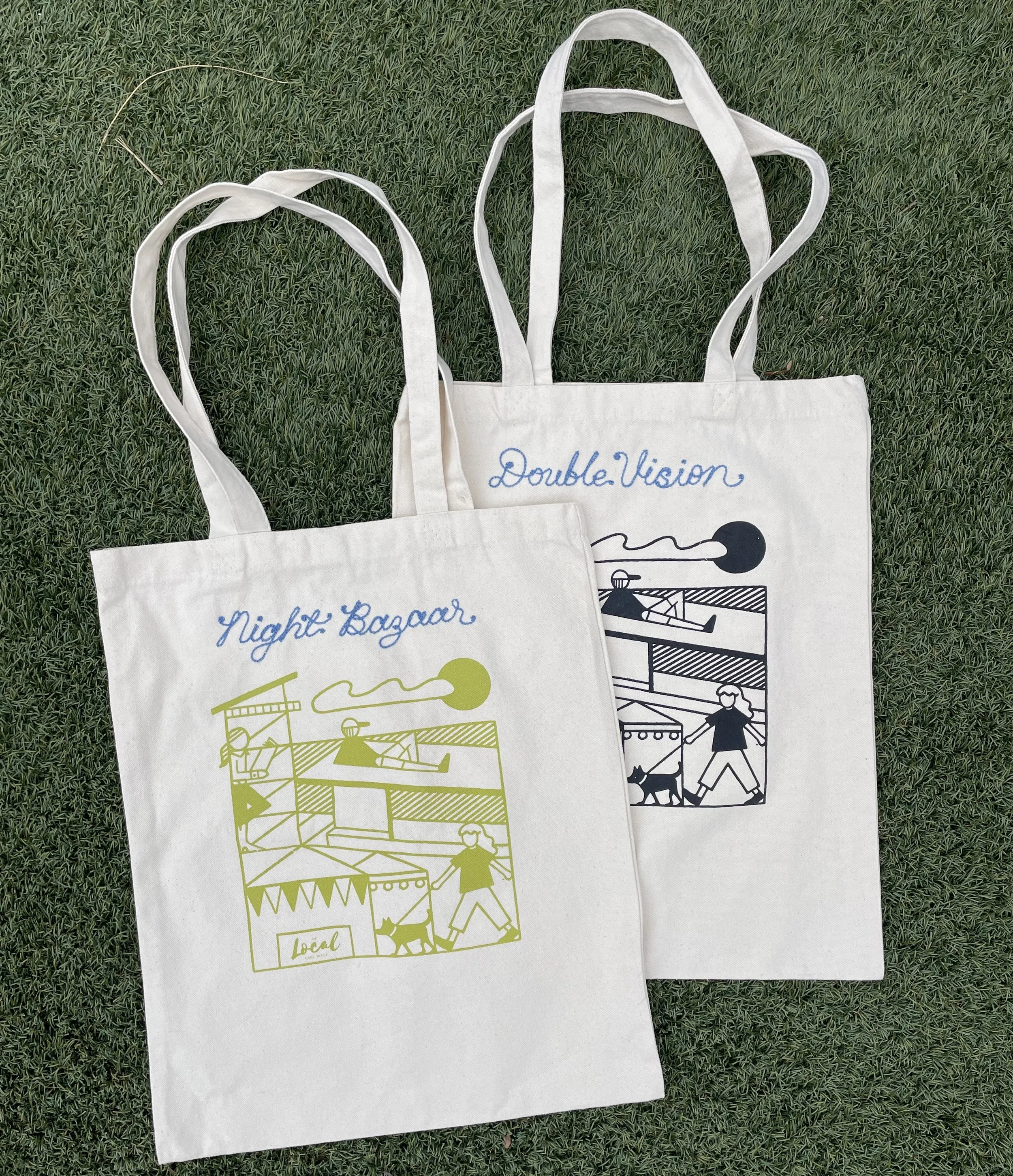 Two cloth tote bags lying on grass, one with green print and the other with black print, both featuring a line drawing of a market scene with a person walking a dog and tents, and embroidered with 'Night Bazaar' and 'Double Vision' respectively.