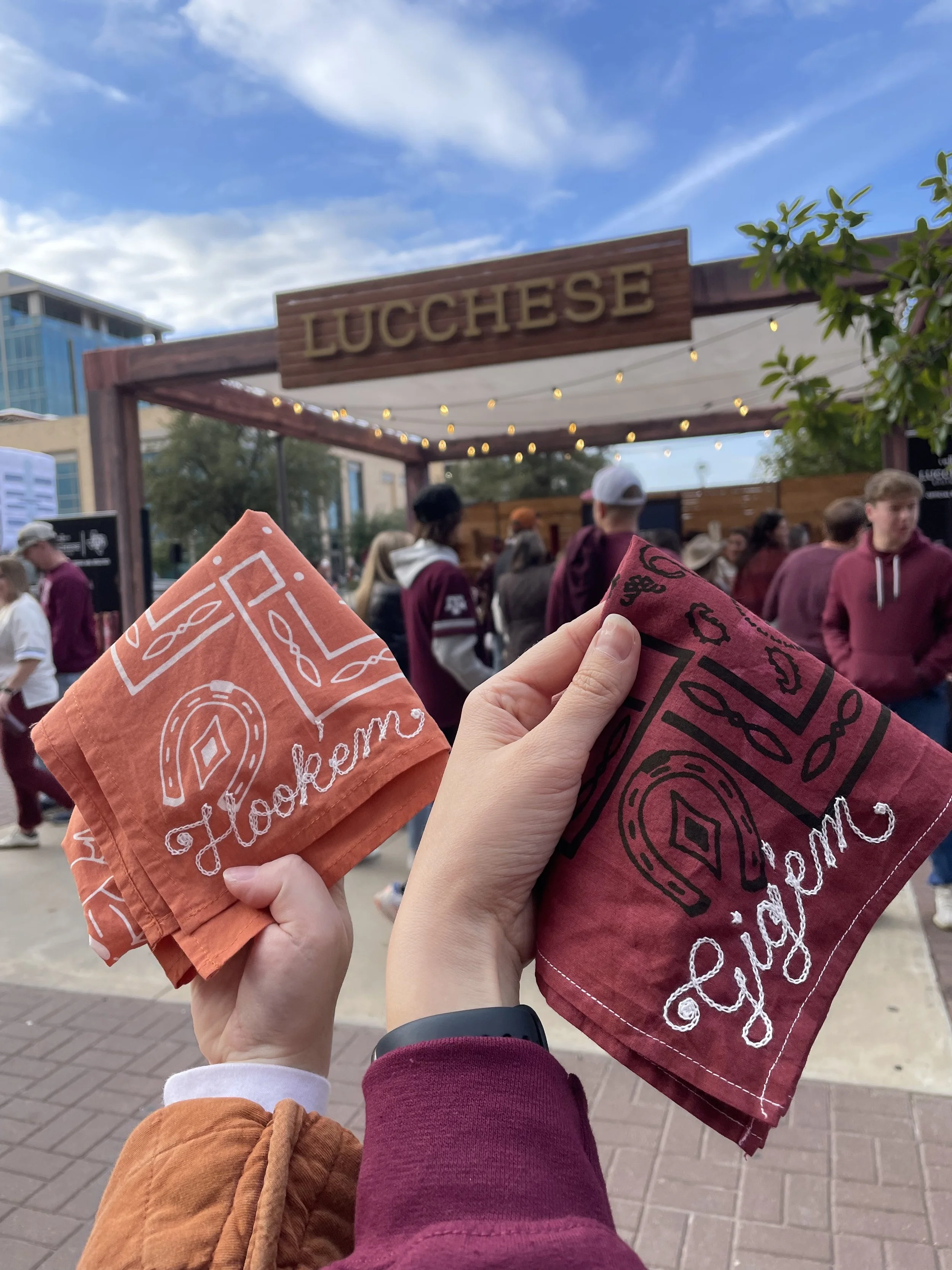 Person holding two embroidered bandanas in front of a booth with a sign that says "LUCCHESE" and a crowd of people. At Texas A&M University in College Station, TX for the TAMU vs. UT game. Double Vision was on-site to provide live embroidery for foot