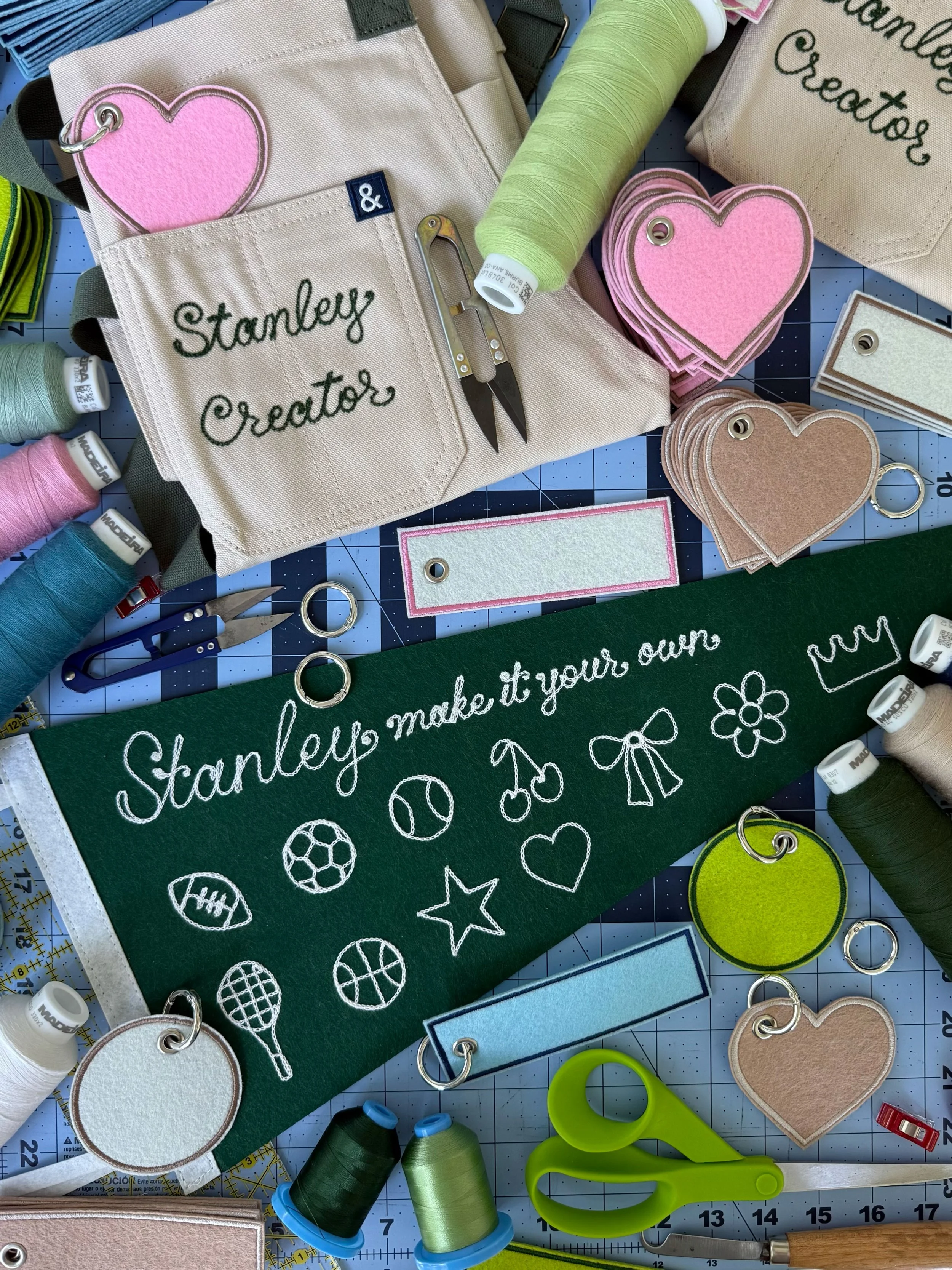 A craft workspace with stitched fabric items, including a green banner that reads "Stanley make it your own" with sports and heart icons, and a beige pouch labeled "Stanley Creator". There are spools of thread in pastel colors, scissors, rings, and various felt cutouts in heart, rectangle, and circle shapes.