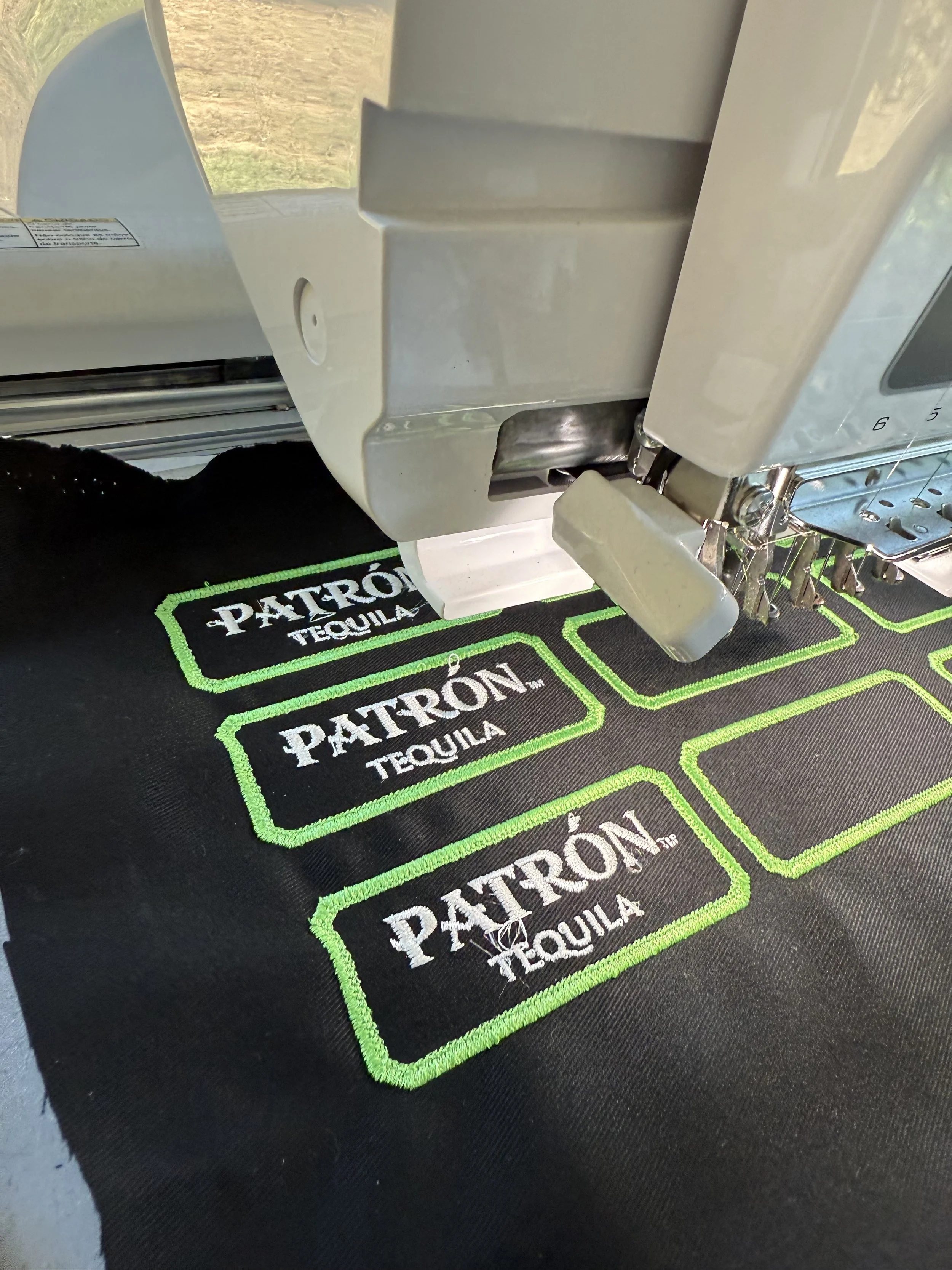 Close-up of embroidery machine stitching patches with the words 'Patrón Tequila' on a black fabric.