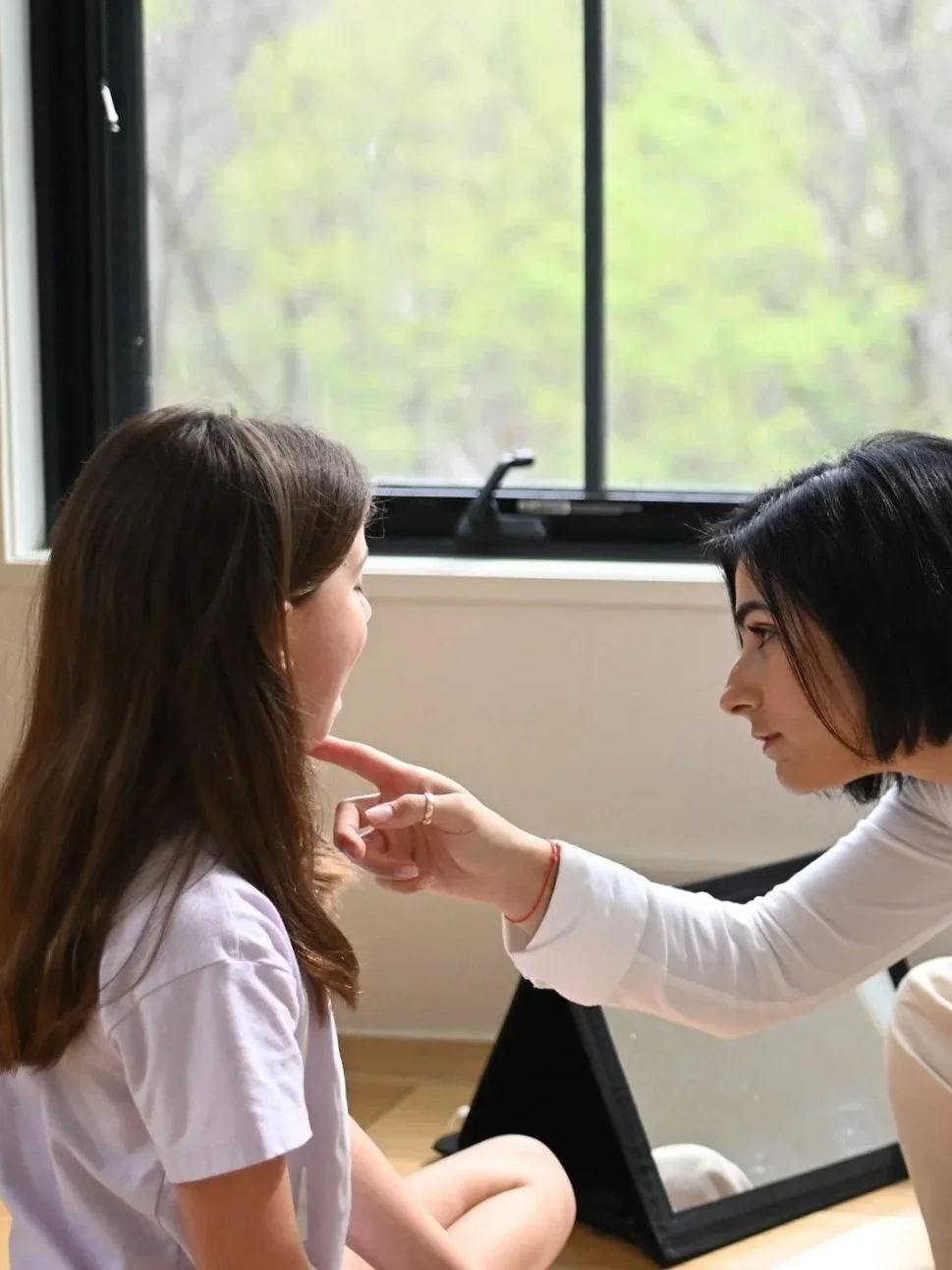 A woman with short black hair examining a young girl with long brown hair, who is seated on the floor in front of a mirror, inside a room near a window with a view of trees.