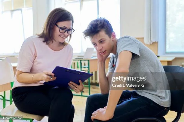 A woman and a young man sitting together in a bright room, looking at a tablet device, smiling, and engaging in conversation.
