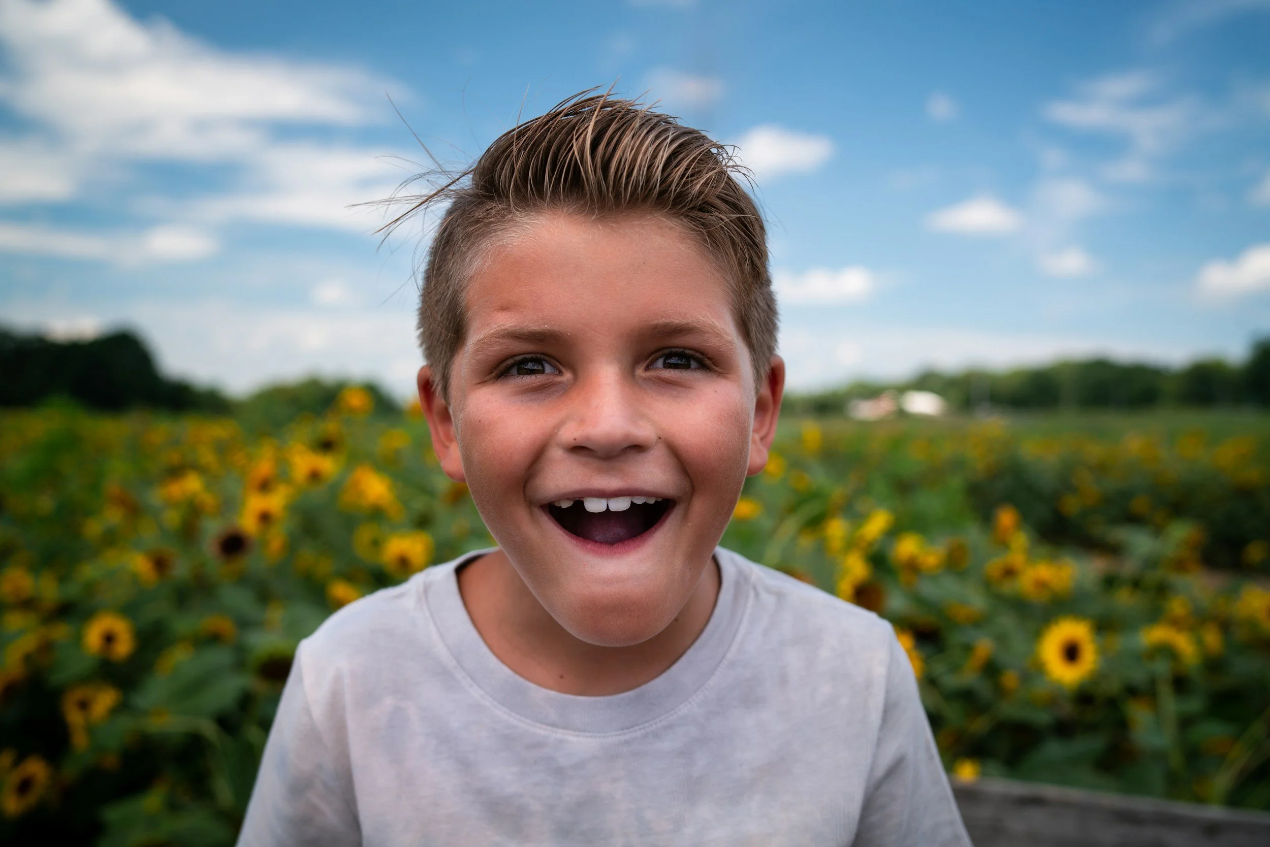 Close-up of a young boy with short, styled hair and a wide, joyful smile, standing outdoors in front of a field of yellow sunflowers with a blue sky and scattered clouds.