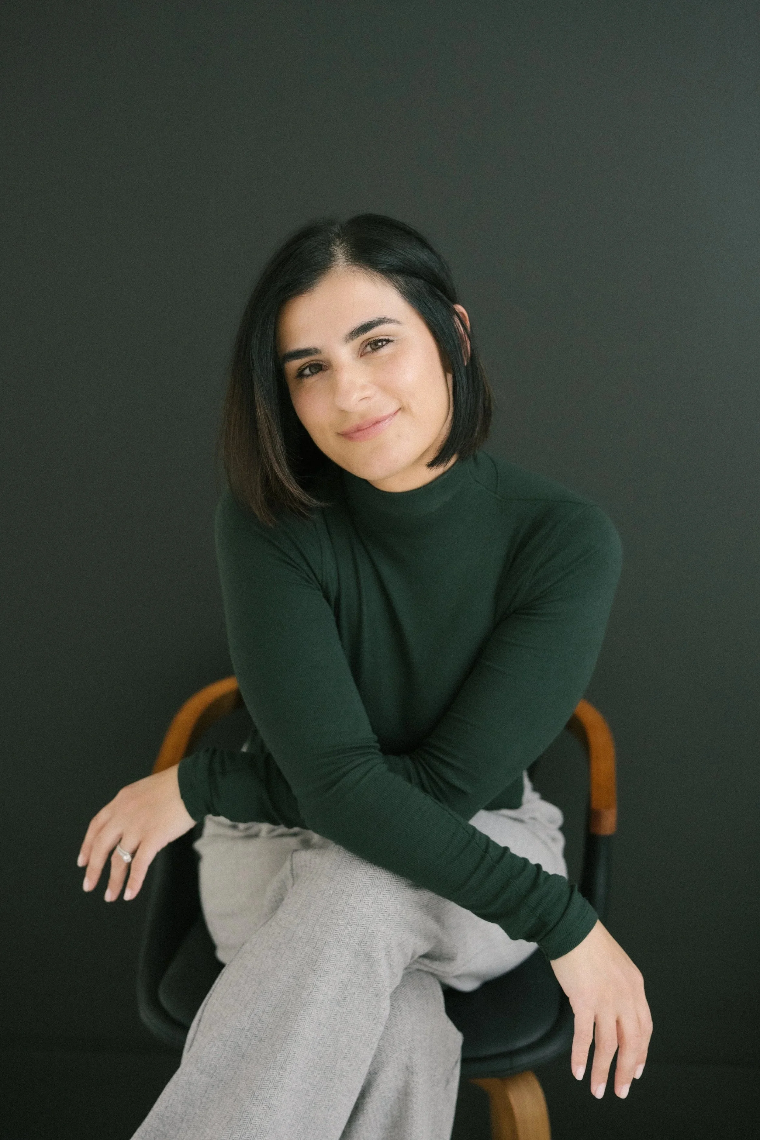 A woman with short black hair, wearing a dark green long-sleeve top and light gray pants, sitting on a black chair with a wooden backrest, against a solid dark background, smiling gently and looking at the camera.