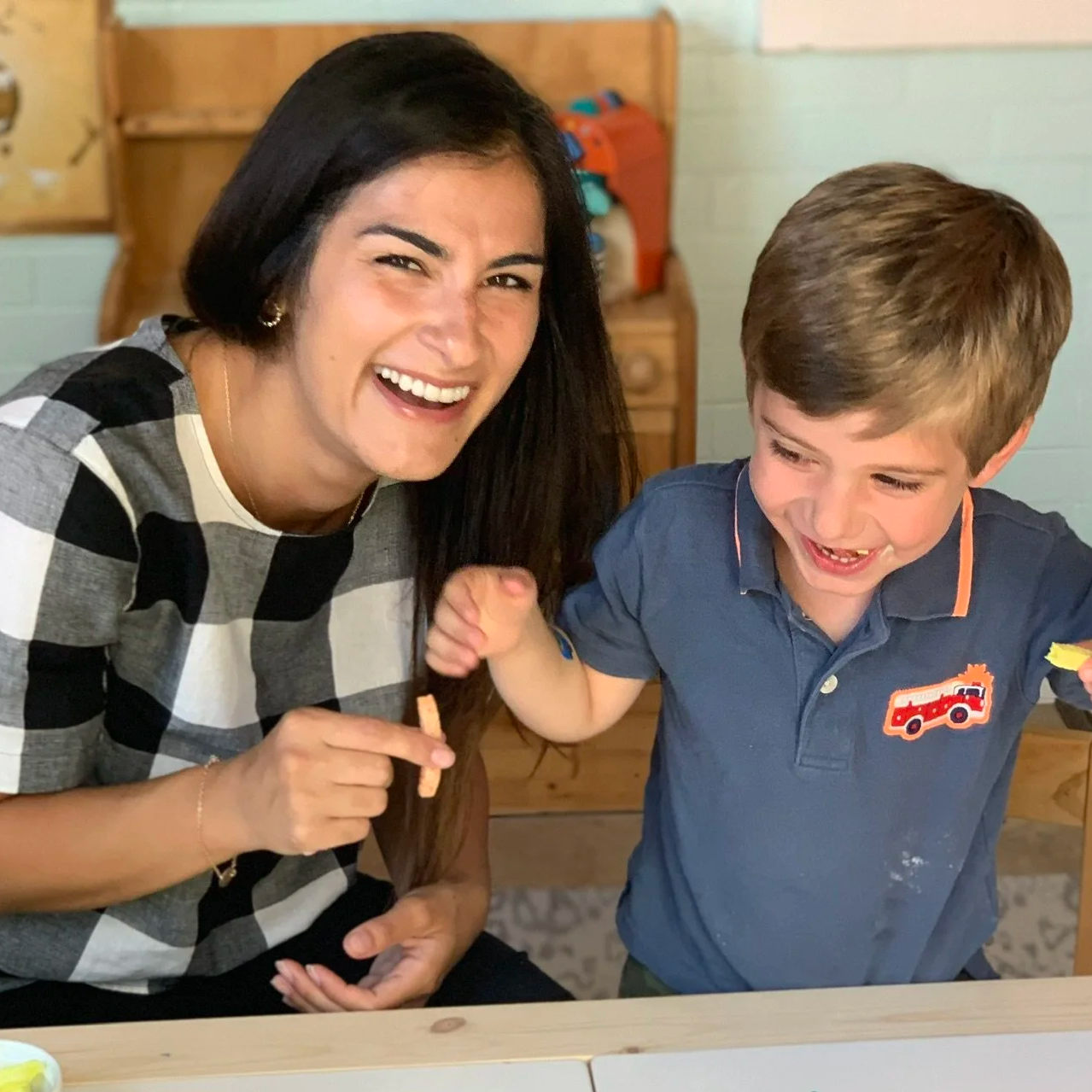 A woman with long dark hair and a checkered shirt smiling and playing with a young boy with brown hair wearing a blue polo shirt with a fire truck emblem, inside a room with wooden furniture in the background.