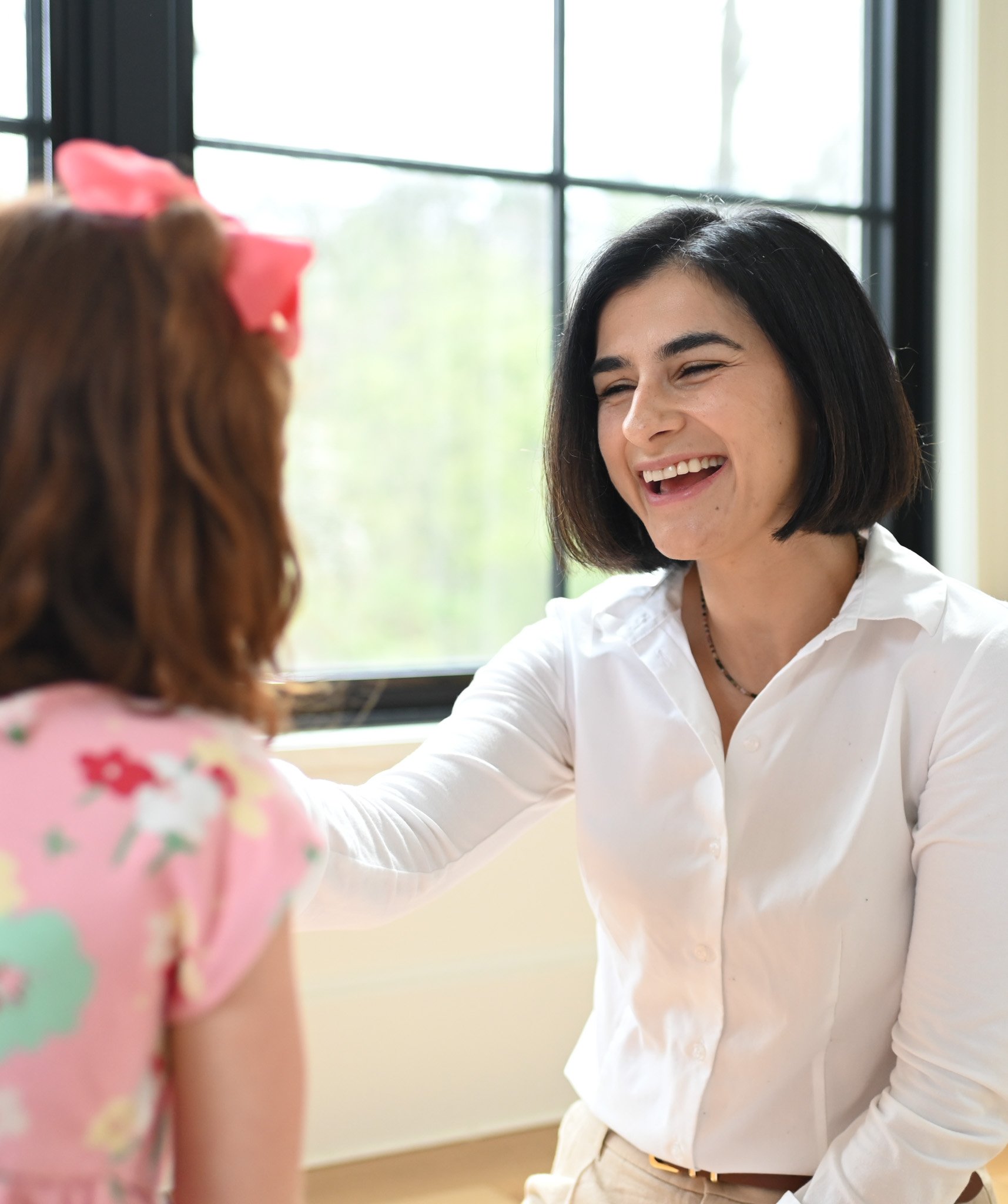 A woman with short black hair smiling and laughing while wearing a white shirt, with a young girl with brown hair and a pink bow in the foreground, outside near a window.