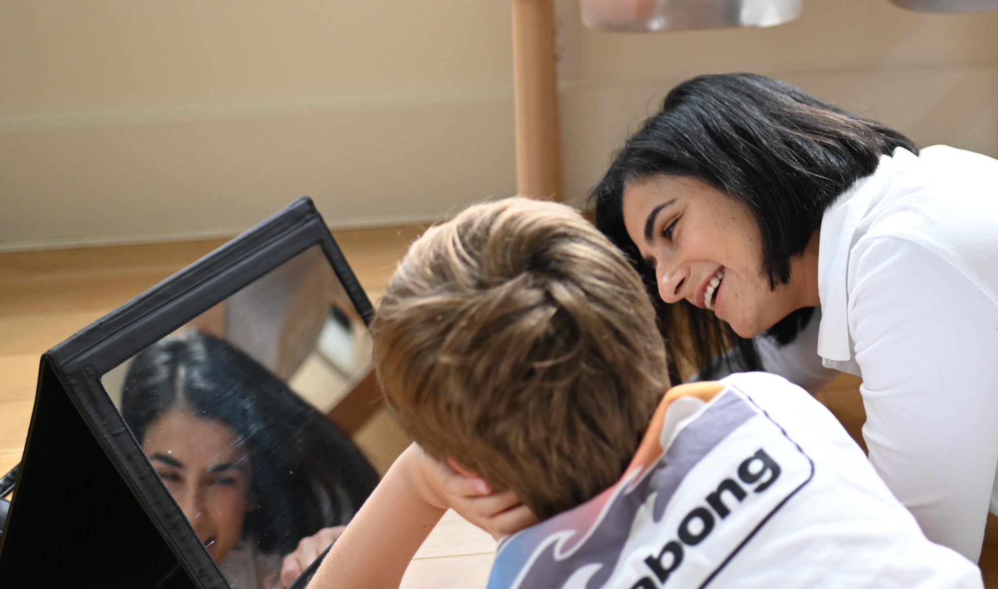 Two children and a woman smiling and touching their heads together in a close-up view, with a laptop and a mirror visible on the table.