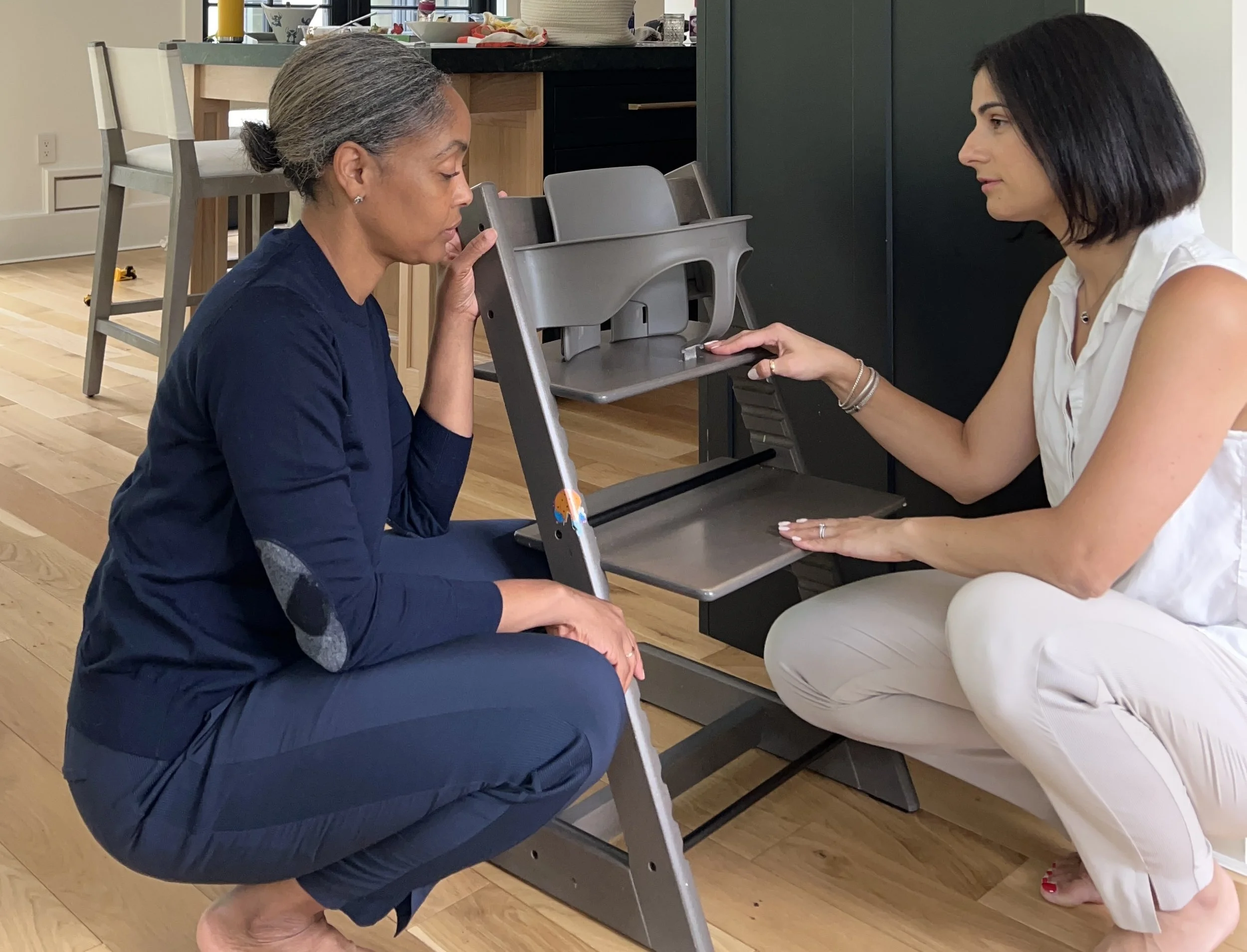 An elderly woman with gray hair tied back, sitting on her ankles, supports her head with her left hand, and appears to be paying close attention during a consultation. A younger woman with shoulder-length black hair, dressed in a sleeveless white blouse and white pants, is sitting across from her, holding the elderly woman’s arm with her left hand and holding a device with her right hand. They are seated at a small table in a kitchen or dining area with a high chair and various items in the background.