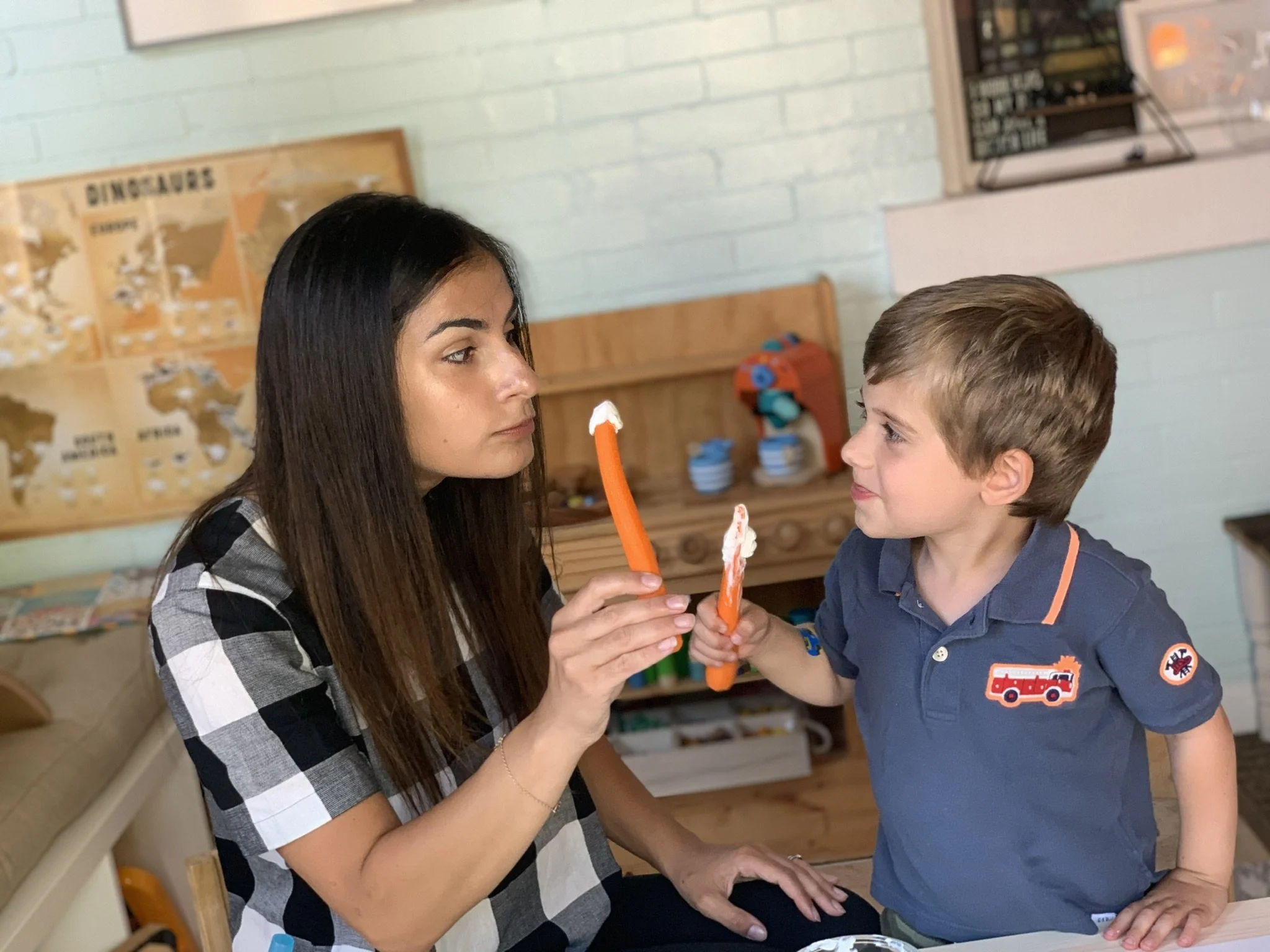 A woman with long dark hair and a young boy with short light brown hair look at each other as they hold orange cleaning sticks in a room decorated with educational posters and a bulletin board.