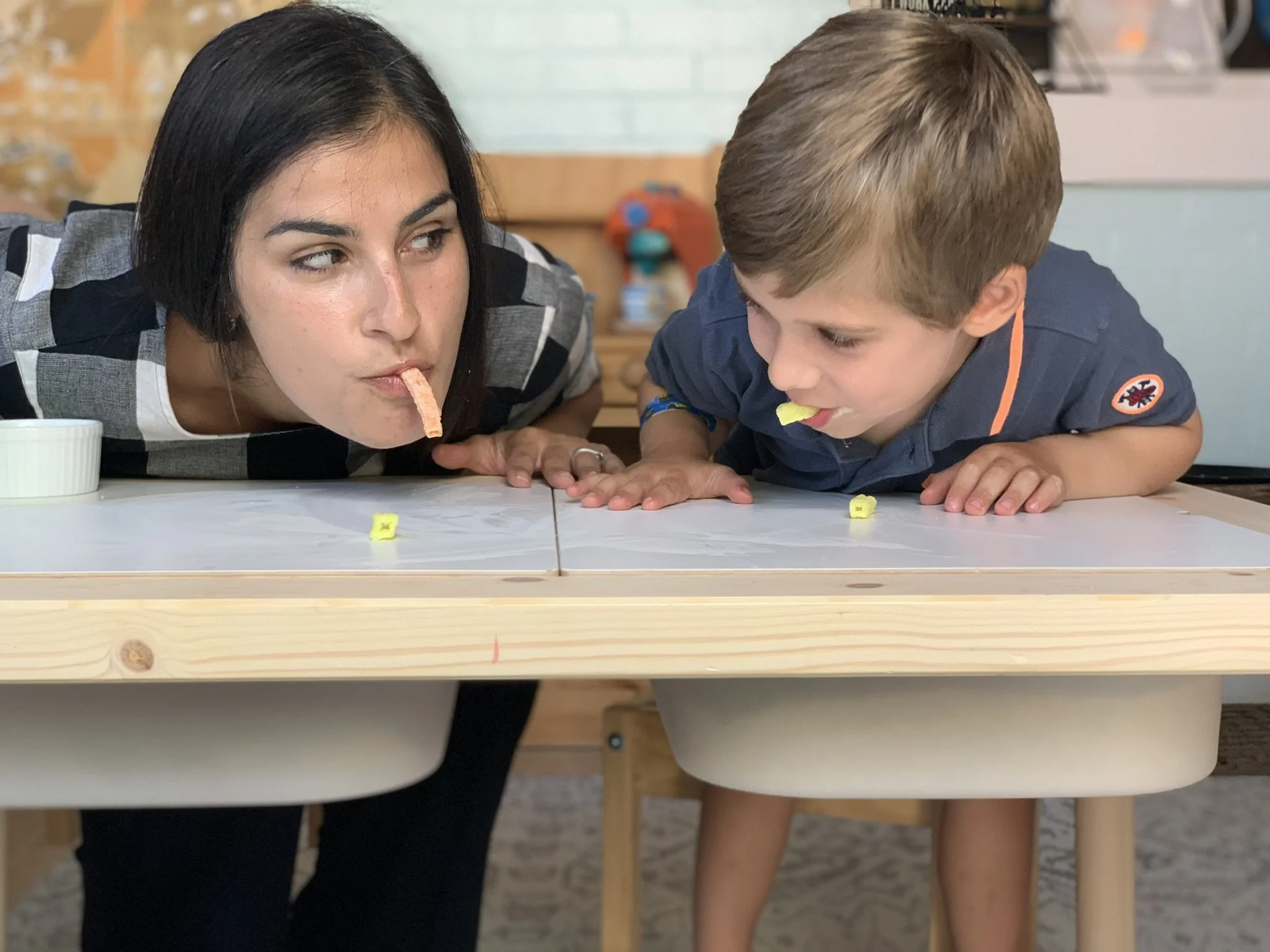 A woman and a young boy are leaning over a table, each with a piece of yellow candy or snack in their mouths, appearing to be engaged in a playful activity or game.