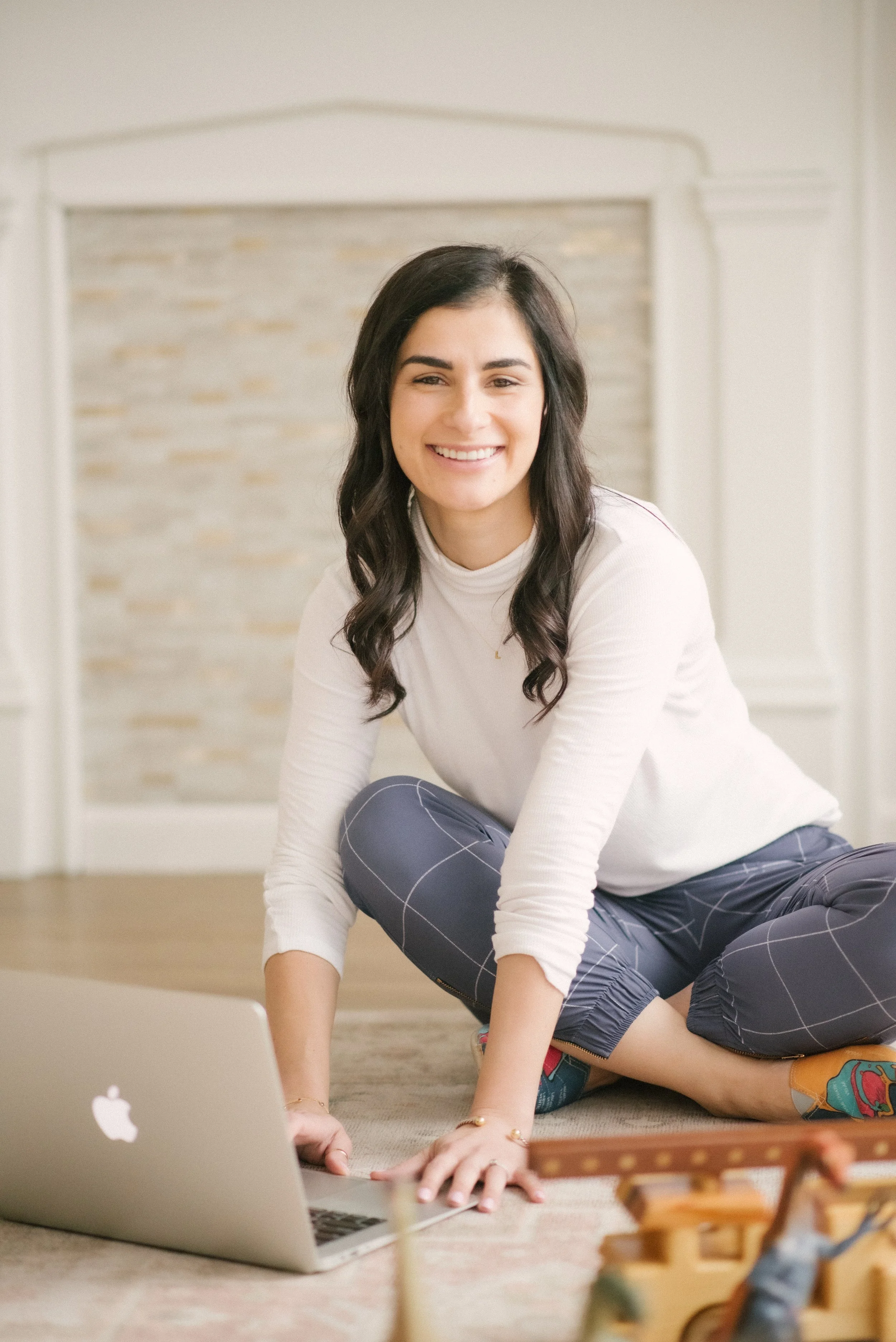 A young woman with dark wavy hair smiling while sitting on the floor in front of a laptop, engaged with her work or activity. She is wearing a light-colored long-sleeve shirt and patterned pants. The background features a light-colored brick wall and a calm, cozy environment.