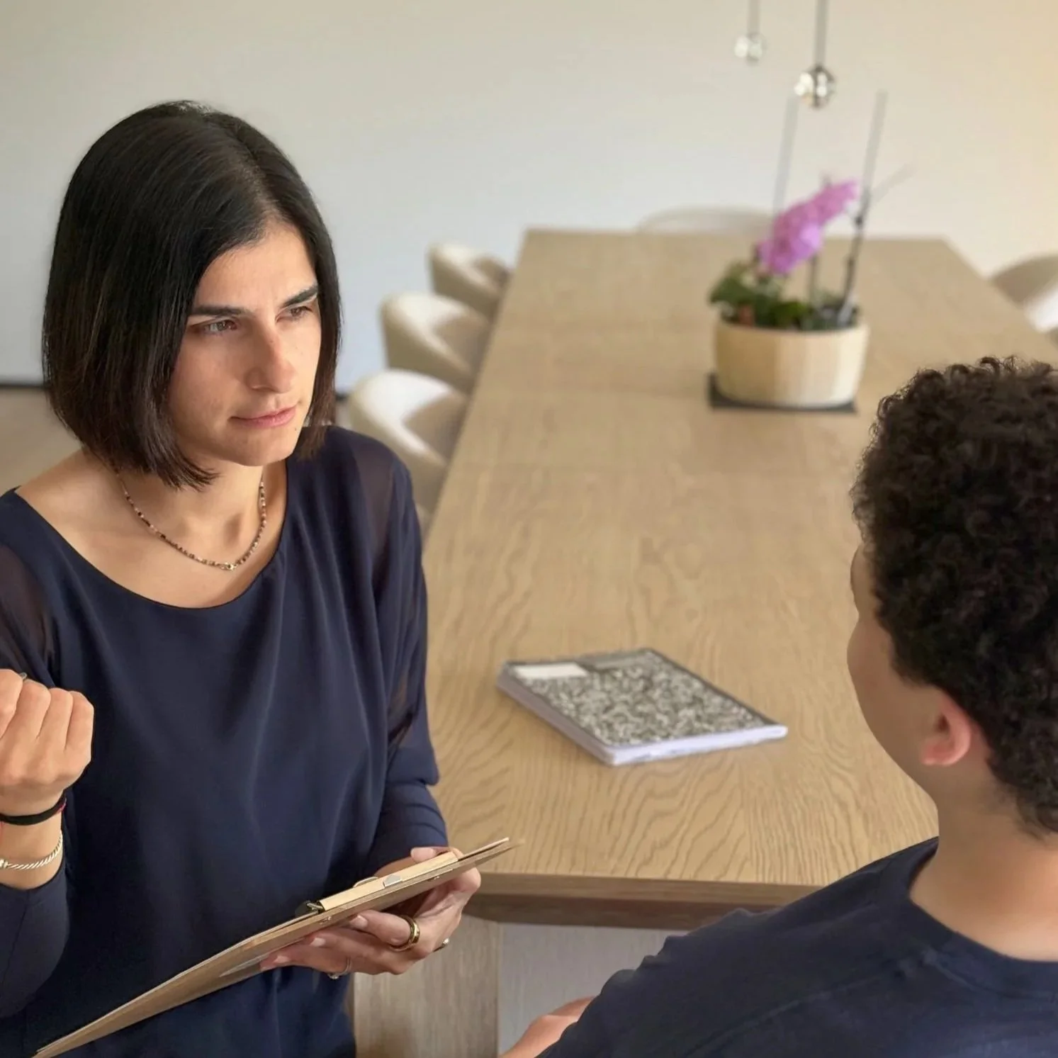 A woman with dark hair wearing a black top and a necklace is holding a notebook and talking to a young boy with curly hair in a black shirt. They are in a room with a long wooden table and a potted purple orchid on the table.