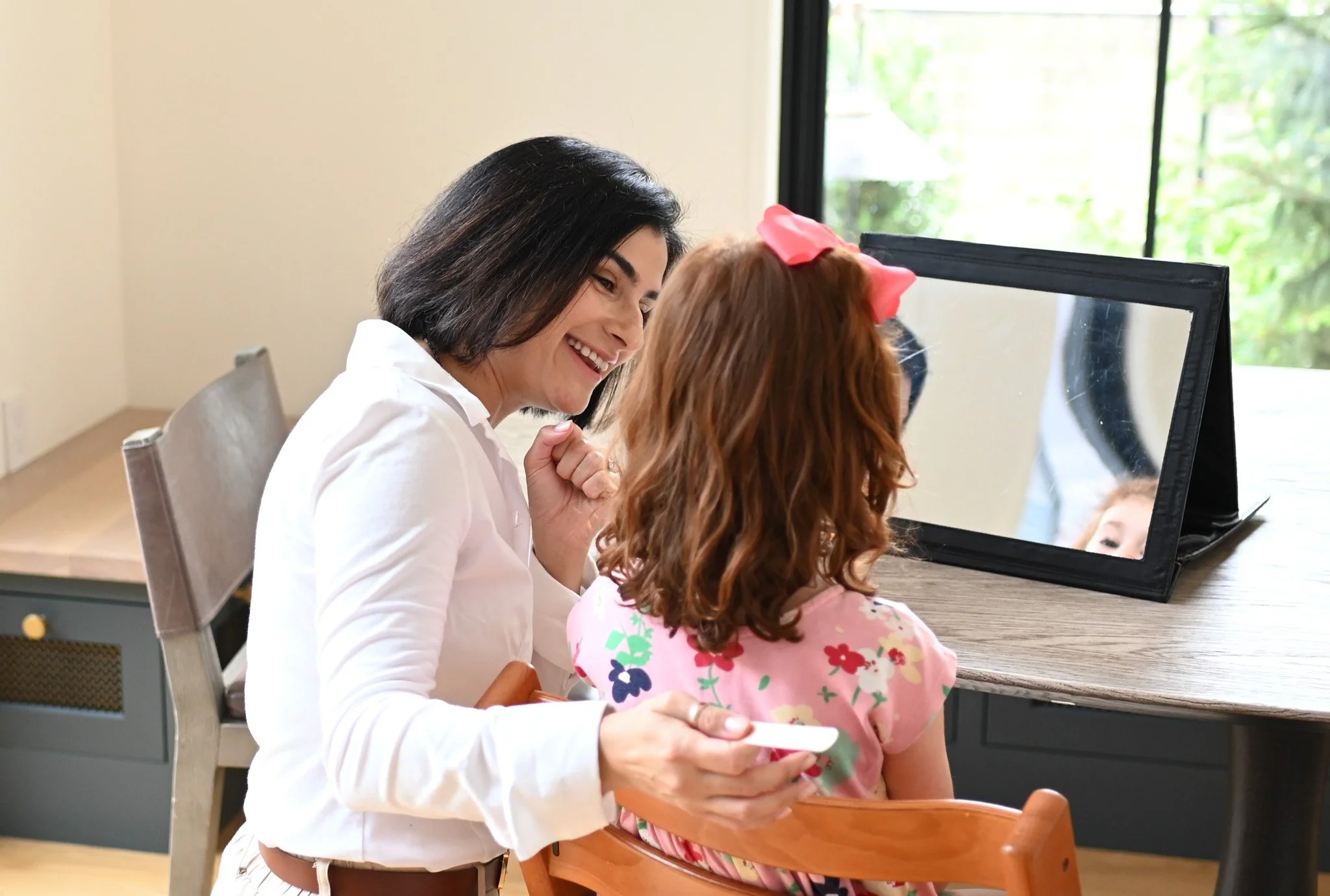 A woman smiling at a girl with curly hair, who is sitting in a high chair at a table, looking in a mirror. The woman is holding a small object, possibly a remote, in her hand. There is a large window in the background with trees outside.