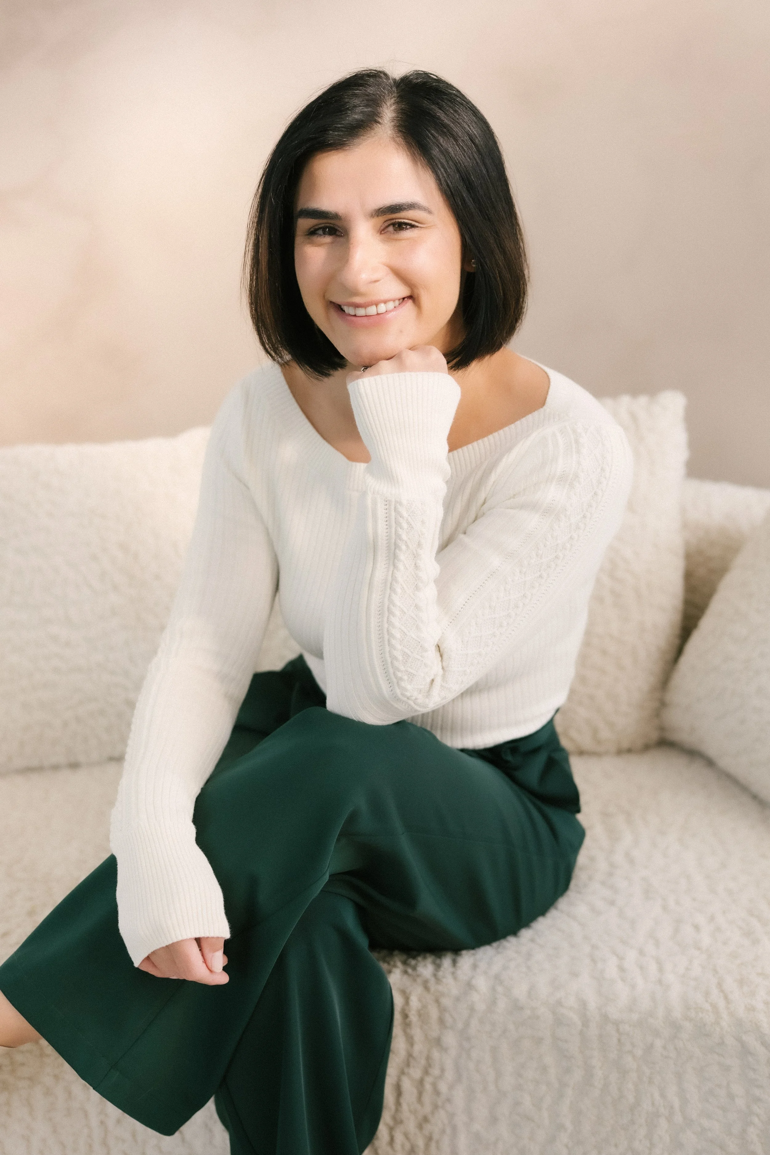 A woman with black hair in a chin-length bob, smiling while sitting on a cream-colored sofa with textured cushions, wearing a white sweater and dark green pants.
