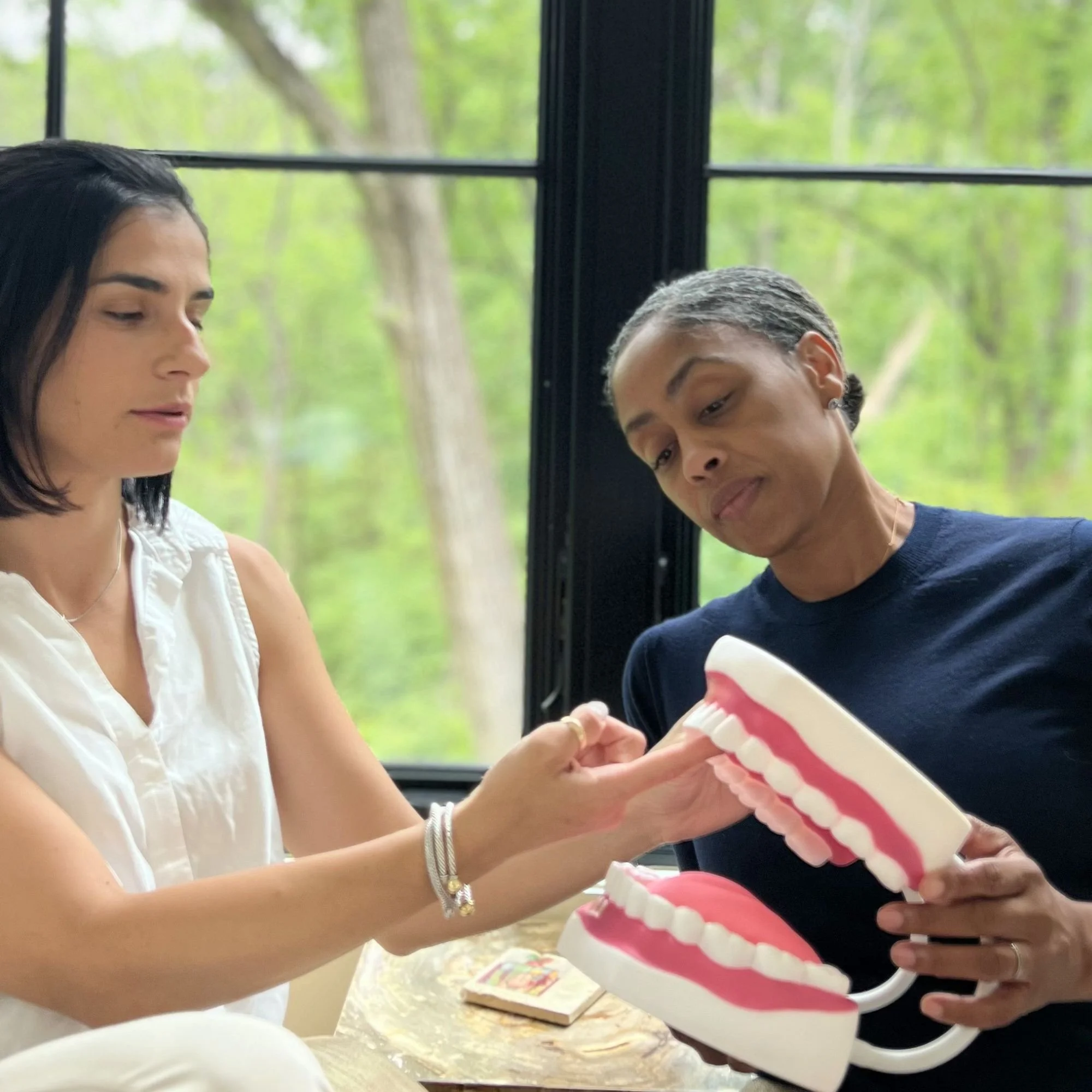 Two women are sitting at a table, sharing a piece of a pink and white frosted cake with sprinkles. The background shows a window with green trees outside.