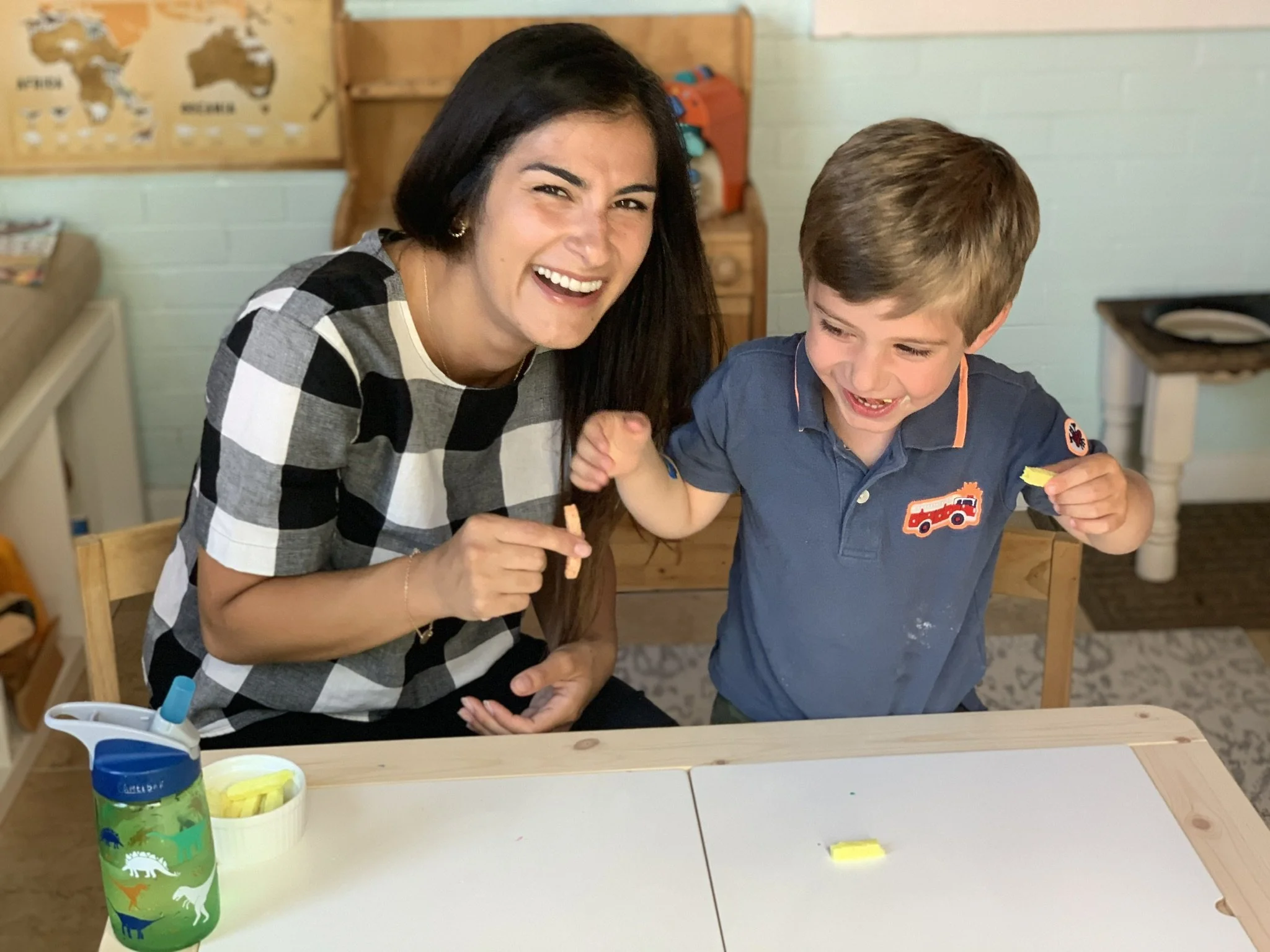A woman and a young boy sitting at a table, smiling and playing with yellow building blocks. The woman has long dark hair and is wearing a black and white checkered shirt. The boy has short brown hair and is wearing a navy blue shirt with a fire truck logo. There are yellow building blocks and a green water bottle with dinosaur illustrations on the table. The background shows a light green wall, a world map, and some shelves with toys.