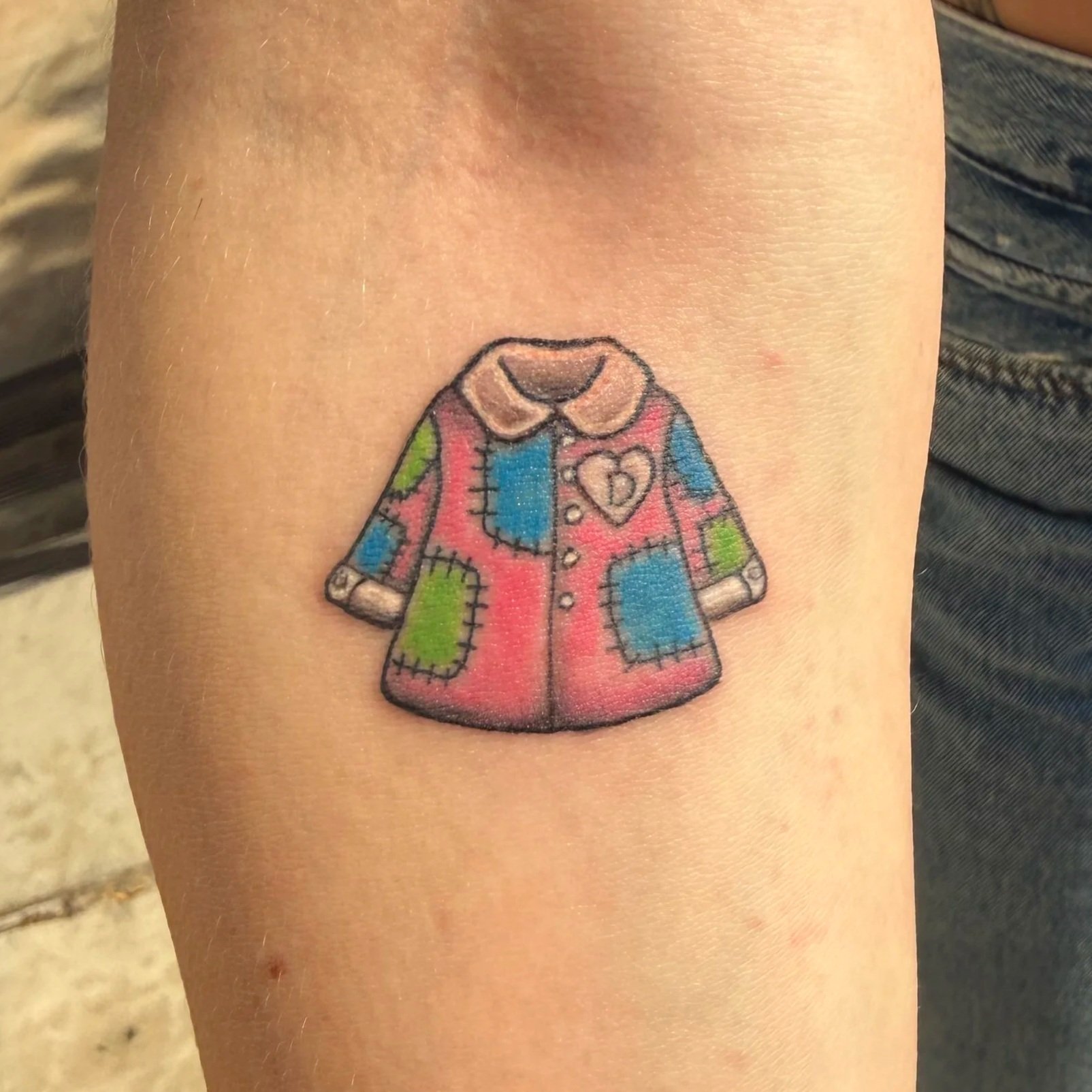 Colorful tattoo of a child's coat with a collar, buttons, and patch pockets on skin Coat of Many Colors tattoo for Dolly Parton Tribute Event. Tattoo by Elizabeth Guerrero at Austin Texas Tattoos in 2026