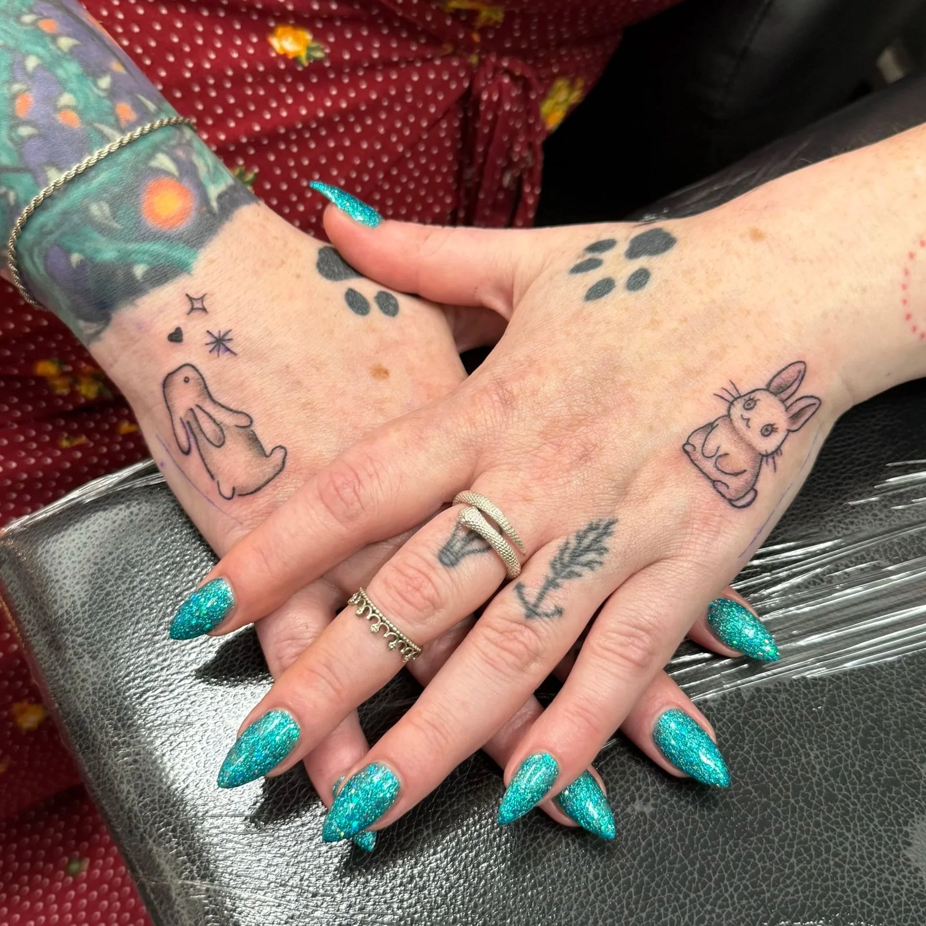 Close-up of two people holding hands with matching tattoos of paw prints and small drawings on their wrists, adorned with colorful glittery nail polish and rings, with a background of a red polka dot shirt and a piece of silver textured material.