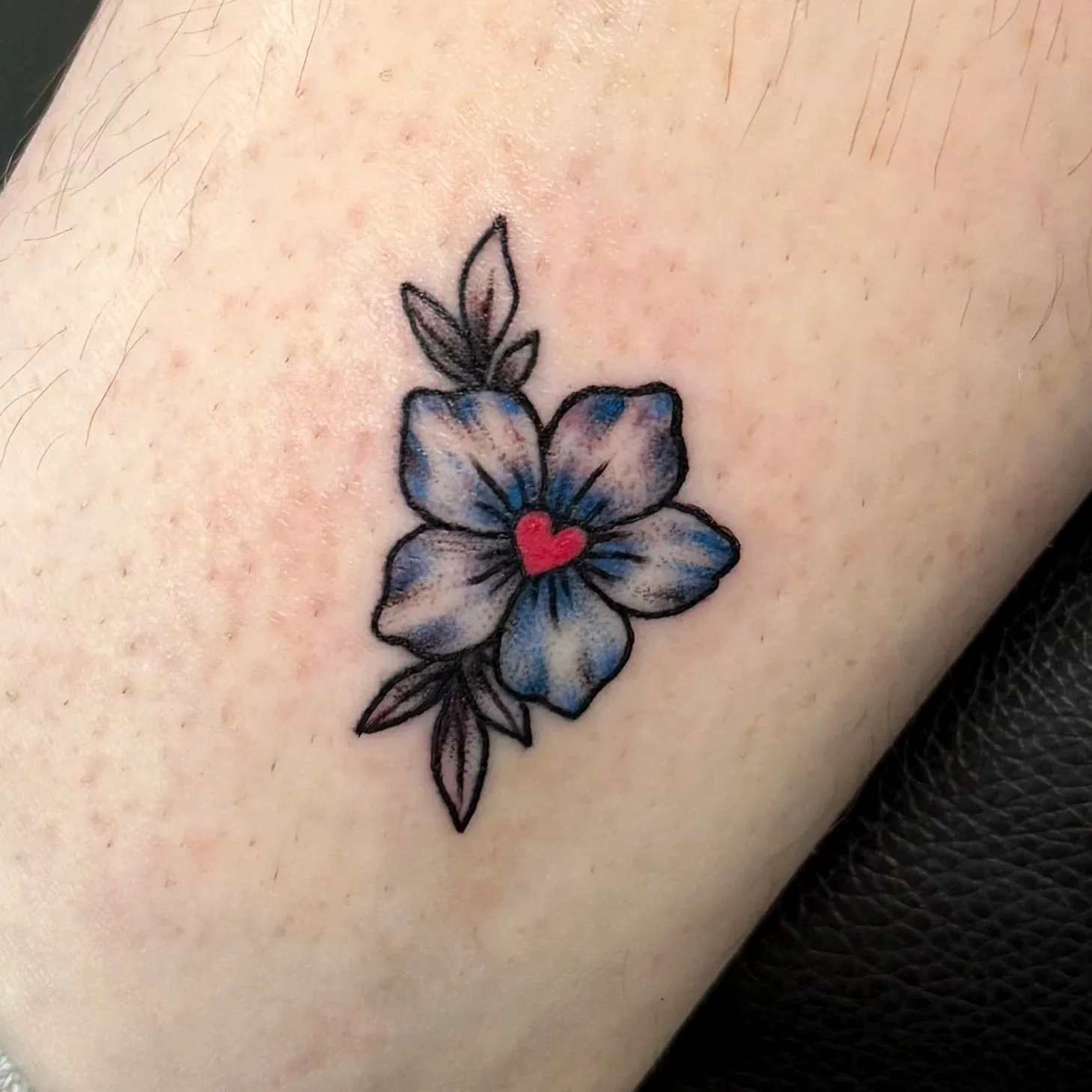 Tattoo of a flower with blue petals and a small red heart in the center on skin. Tattoo by Elizabeth Guerrero at Austin Texas Tattoos in 2026