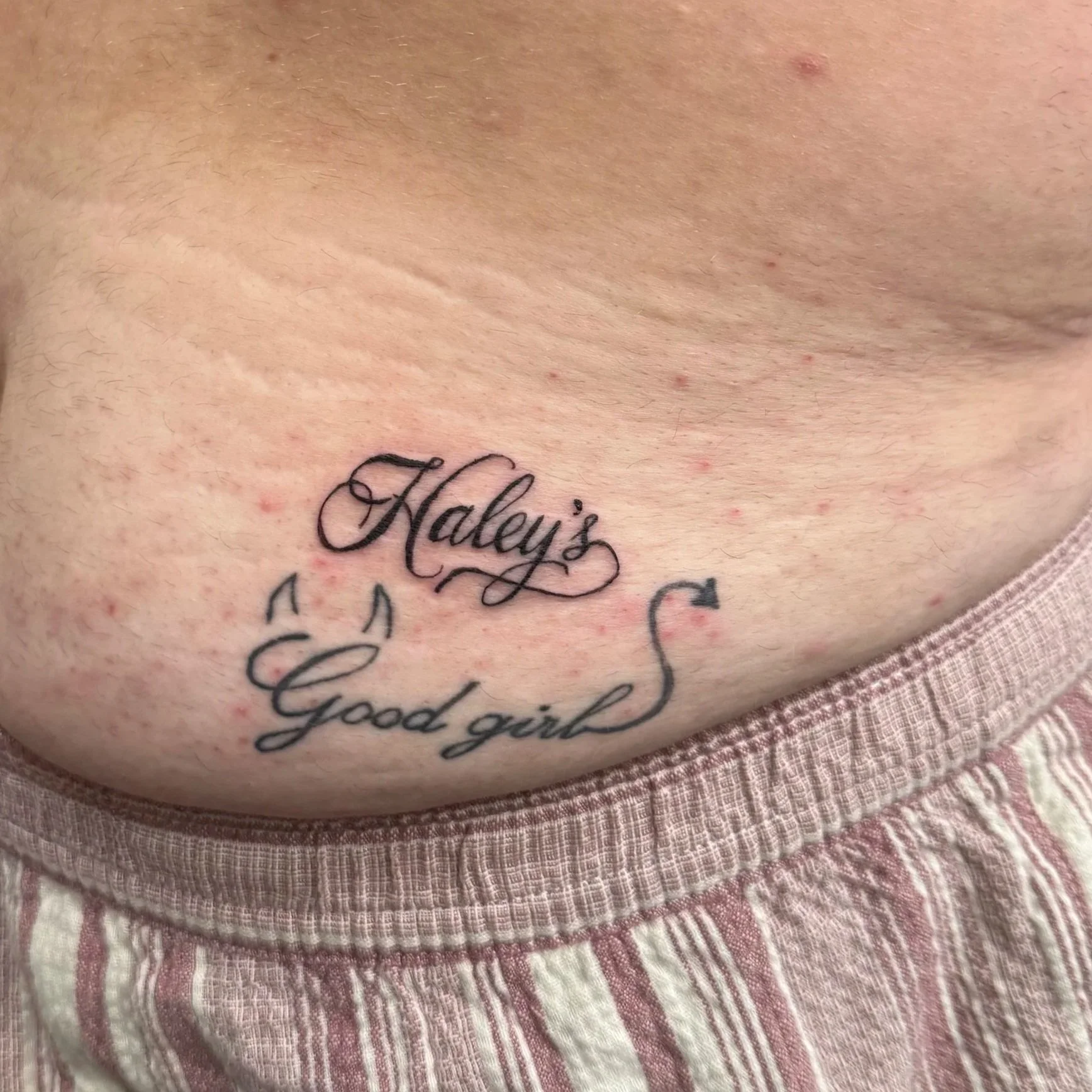 A tattoo on skin with the words 'Haley's' and 'Good girl' written in cursive. The skin has small red blemishes and is partially covered with pinkish fabric.