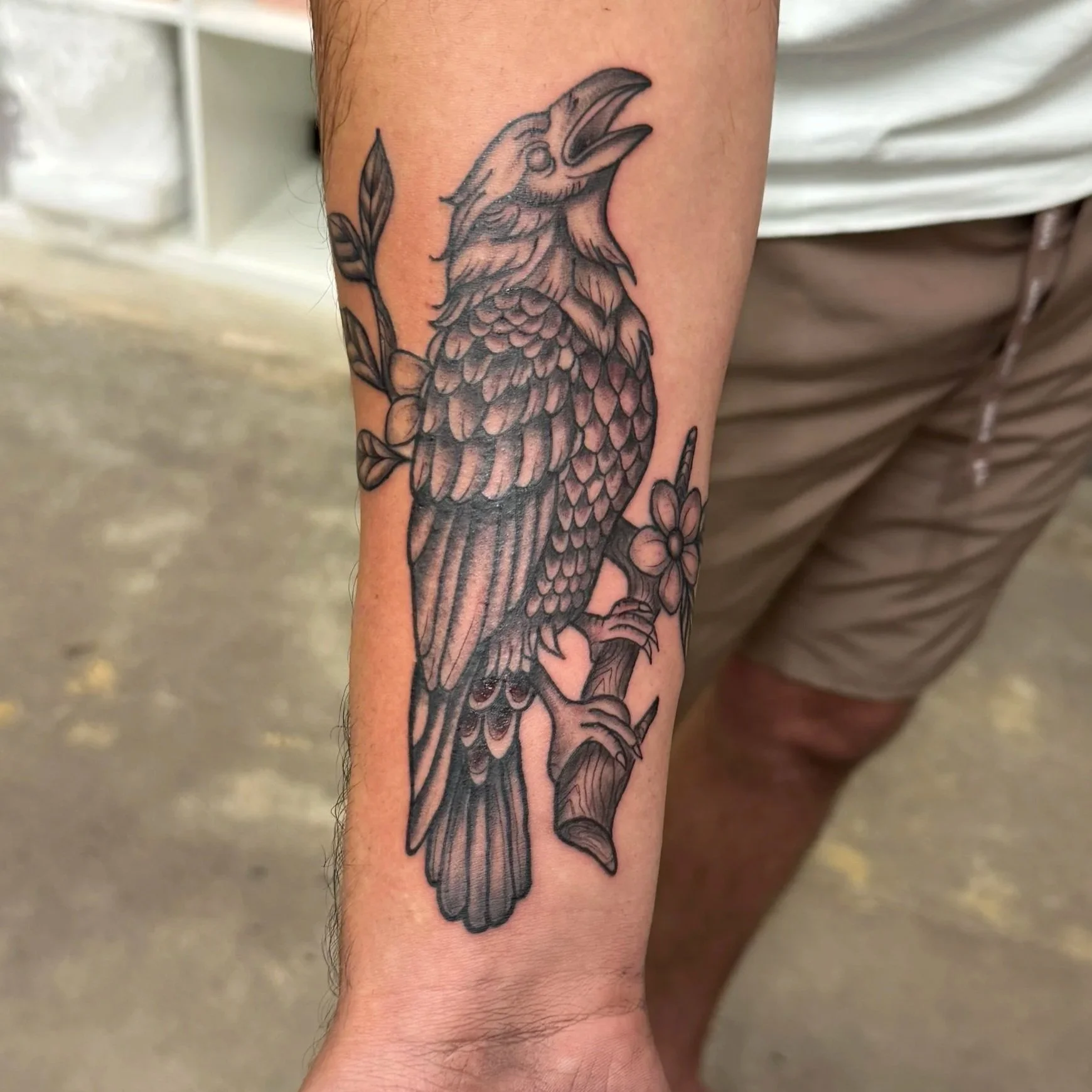 Black and gray tattoo of a bird with its mouth open, surrounded by flowers and leaves on a person's forearm. Tattoo by Elizabeth Guerrero at Austin Texas Tattoos in 2026