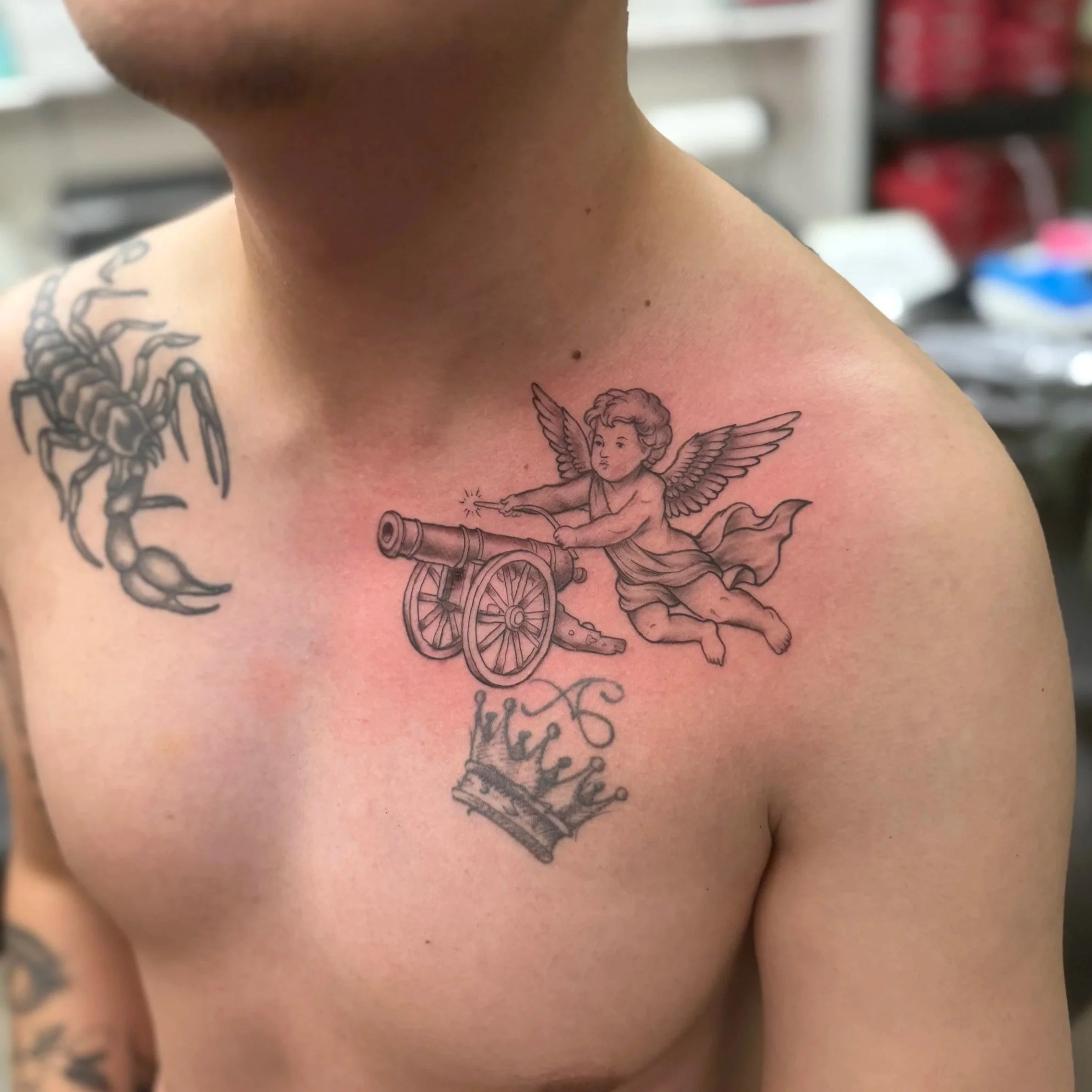 Close-up of a person's shoulder with tattoos, including a cherub, a scorpion, and text, in a setting with blurred objects in the background.