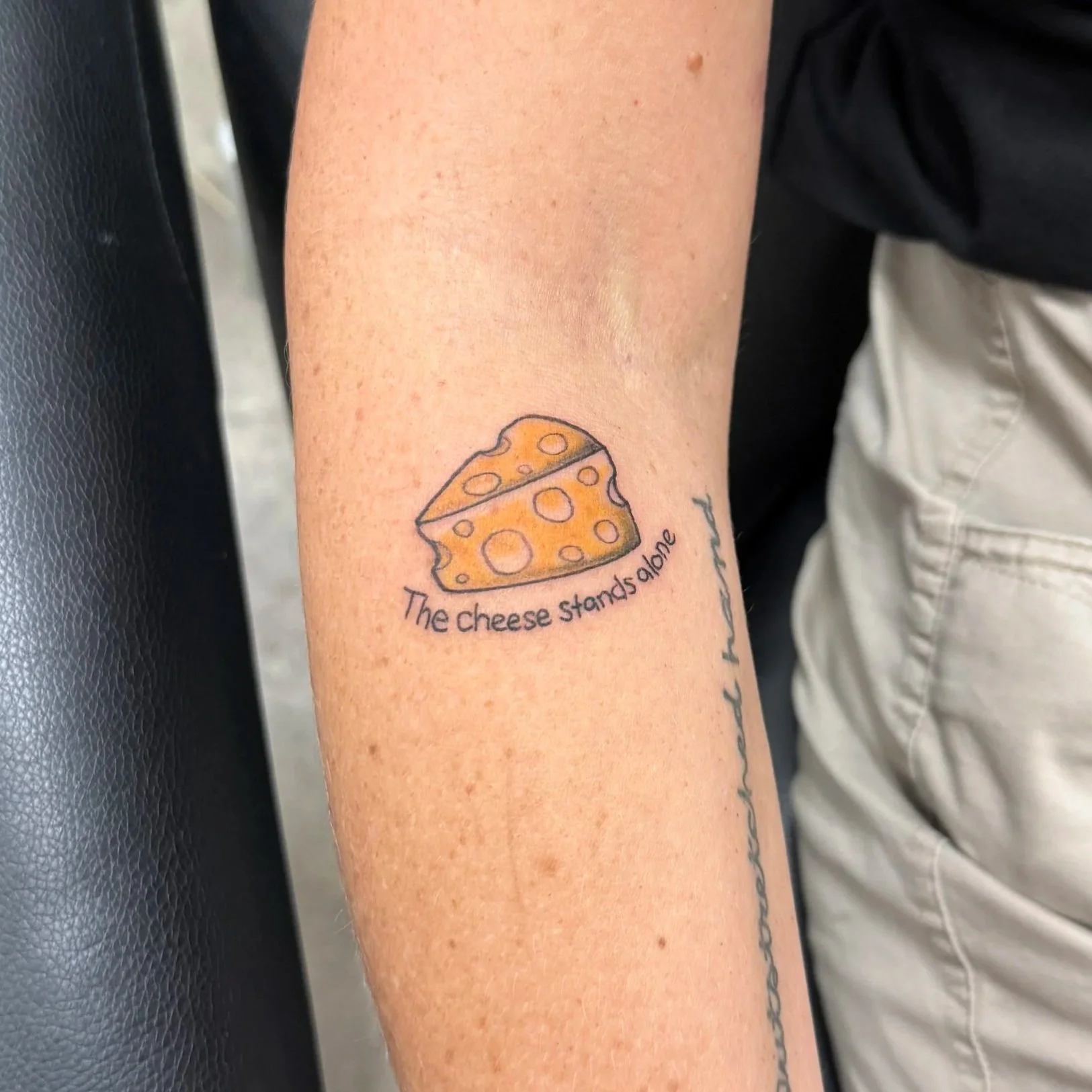 A tattoo of a wedge of cheese with holes, accompanied by the phrase 'The cheese stands alone' on a person's inner forearm.