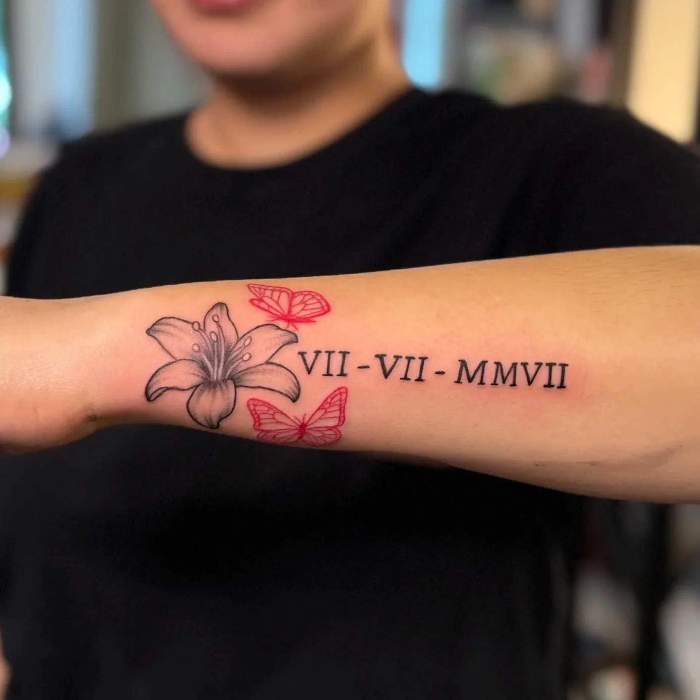 Tattoo on person's forearm featuring a black and gray lily flower, two red butterflies, and Roman numerals VII - VII - MMVII.