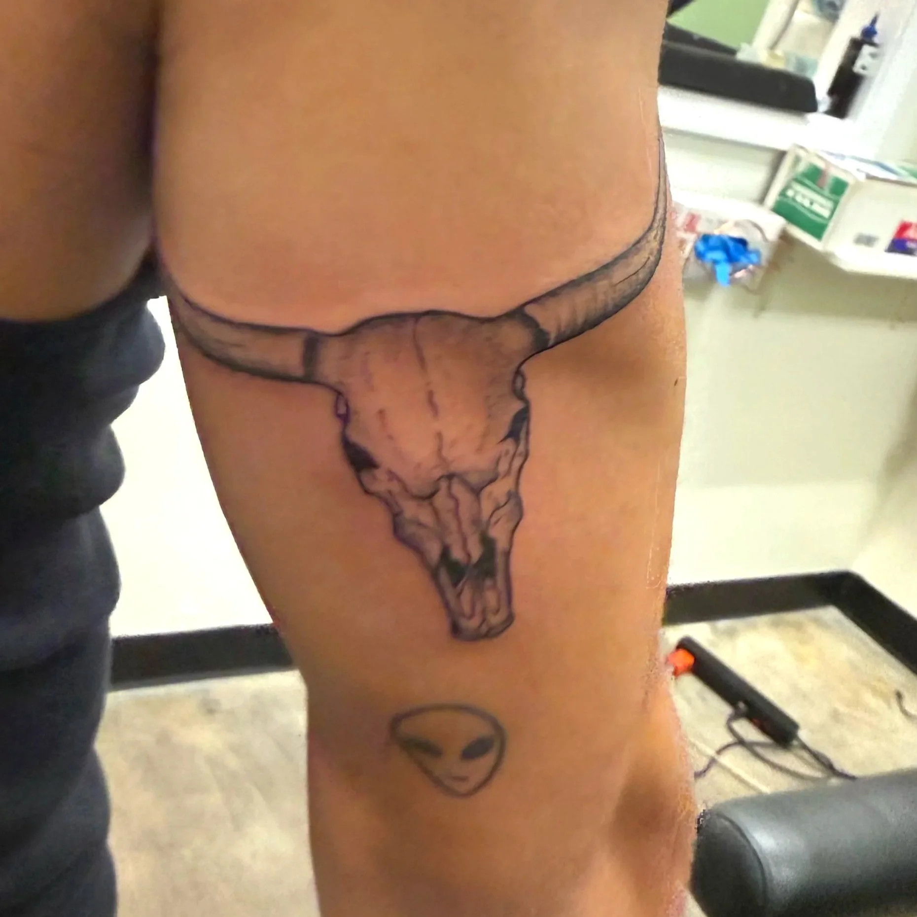 Close-up of a person's lower back and upper buttocks, with black ink tattoo of a snake coiled along the lower back extending downward. The background shows a blurry bathroom setting with a counter, various items, and a box of bandages.