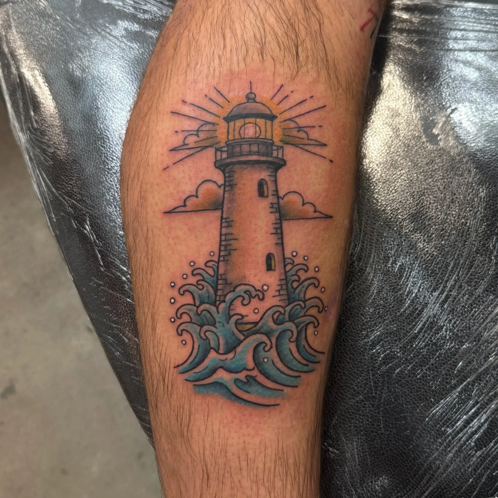 A tattoo of a lighthouse emitting sun rays, with clouds and waves below, on a person's upper arm.