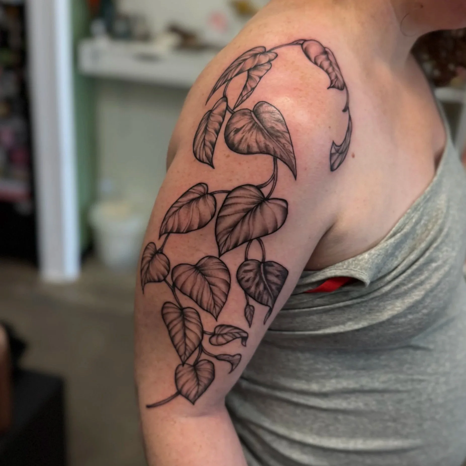 A person's upper arm with a black outline tattoo of a vine with heart-shaped leaves.