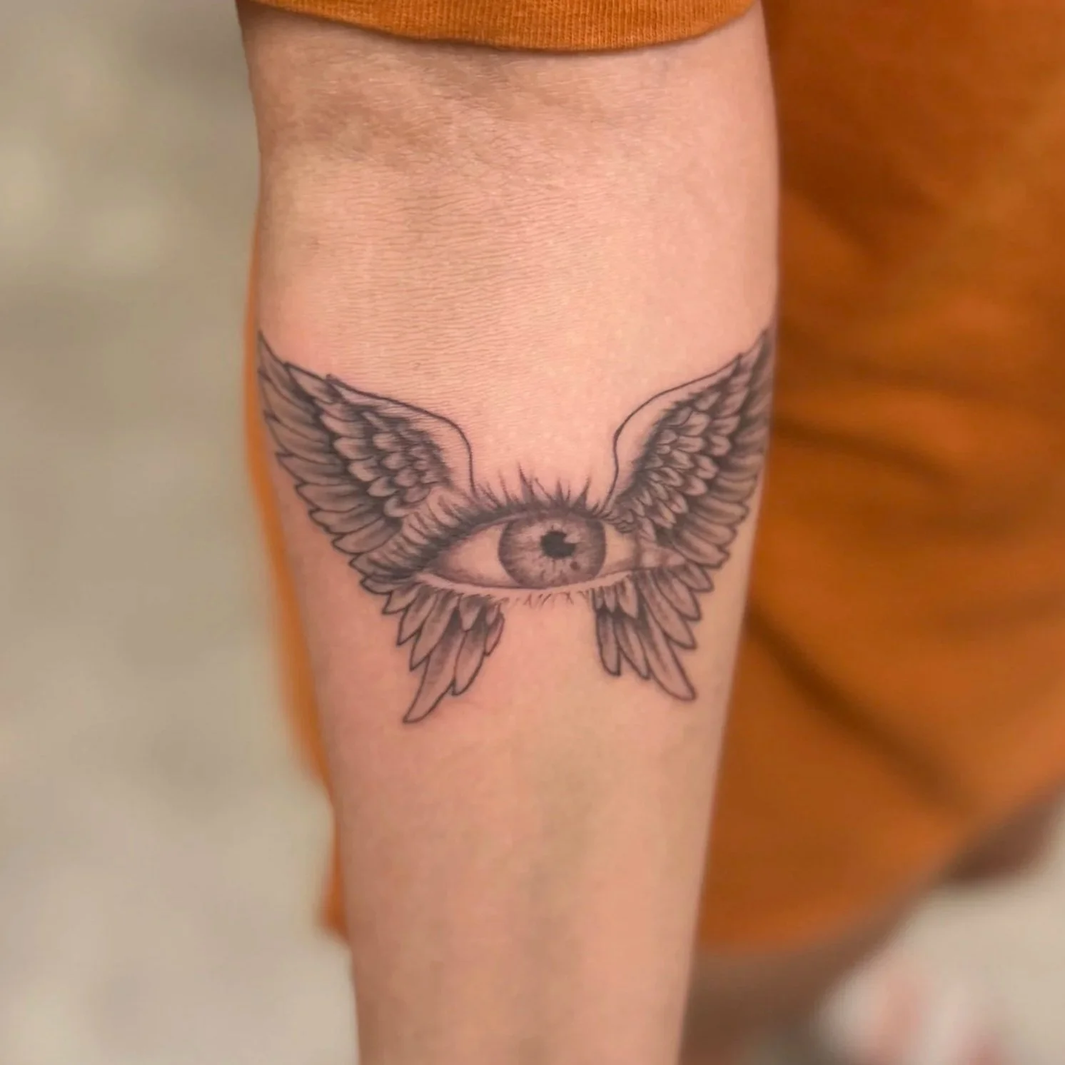 Tattoo of an eye with wings and feathers on upper arm.