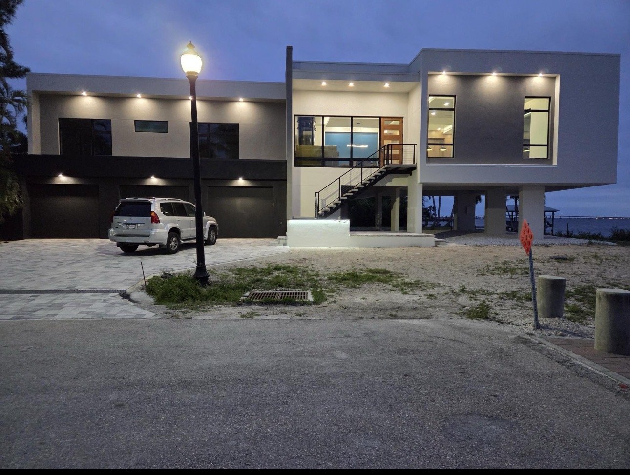 Modern two-story house with large glass windows, a staircase outside, and a garage with a white SUV parked in front, illuminated by exterior lights at dusk.