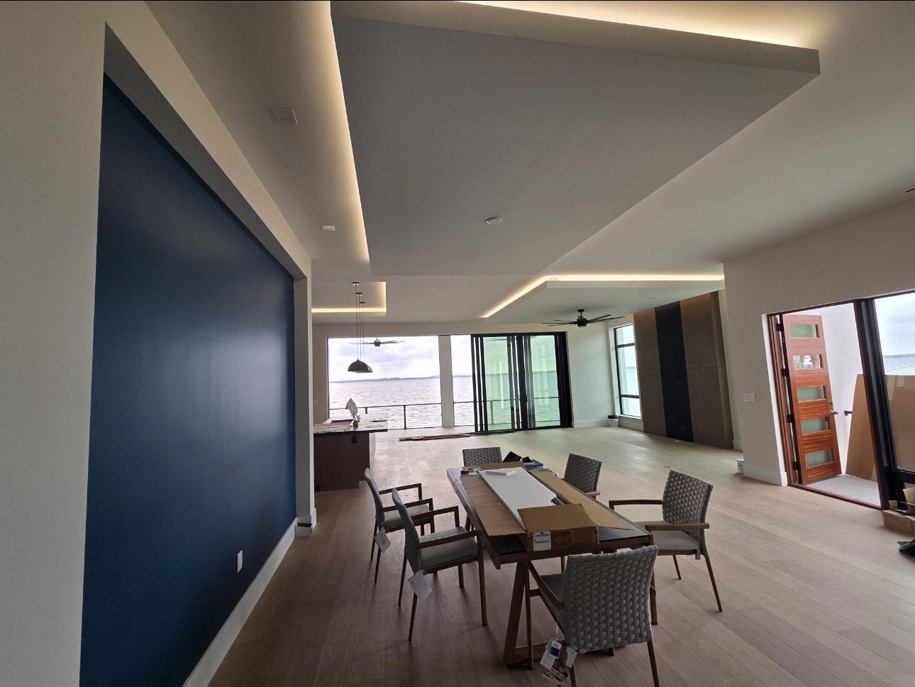 Interior of a modern, spacious living area with large windows overlooking water, a blue accent wall, a dining table with chairs, and doors leading to an outdoor space.