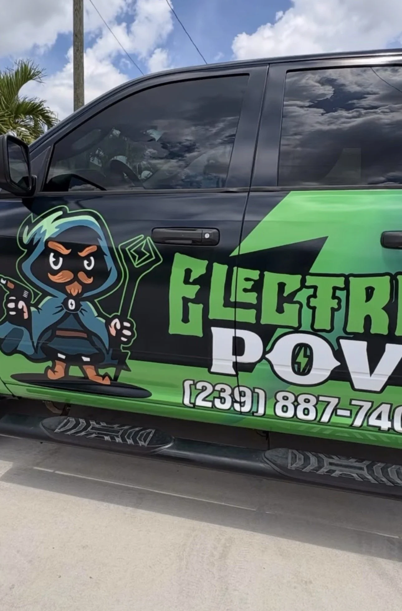A vehicle with a graphic design featuring a cartoon character dressed as a wizard with a mustache, holding a lightning bolt and a smartphone. The vehicle displays the words "Electric Power" and a phone number.